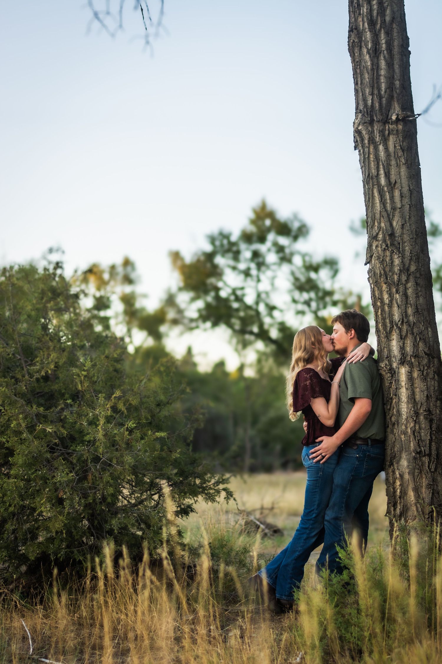 Couple kisses leaning against tree trunk in golden field surrounded by lush green foliage.