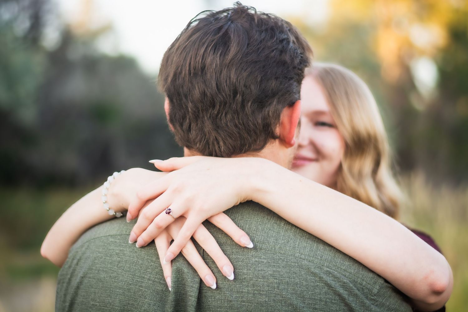 Woman hugging man from behind showing engagement ring, smiling outdoors in natural setting.