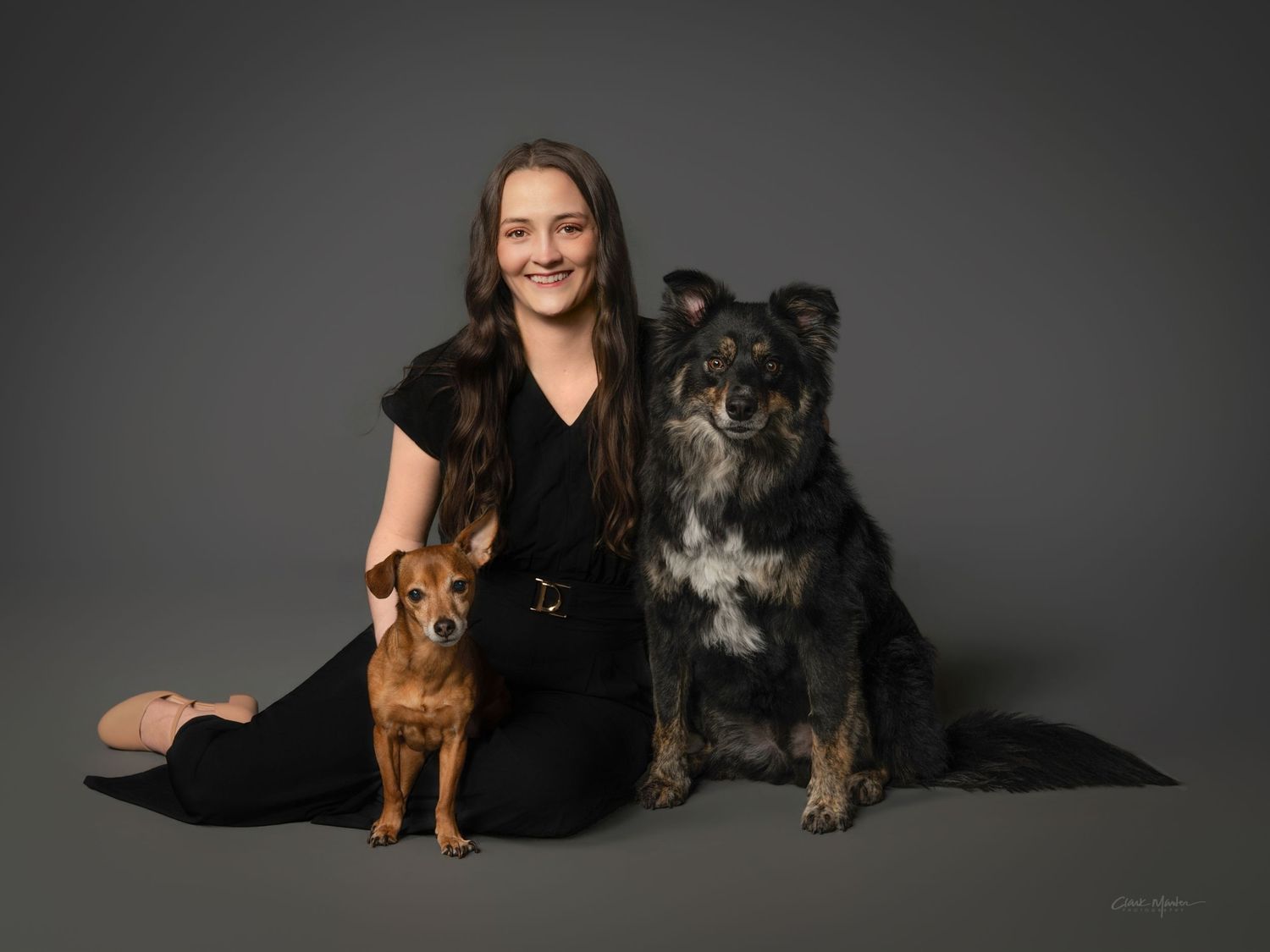 Smiling young woman sitting on floor with two dogs against a gray studio background.