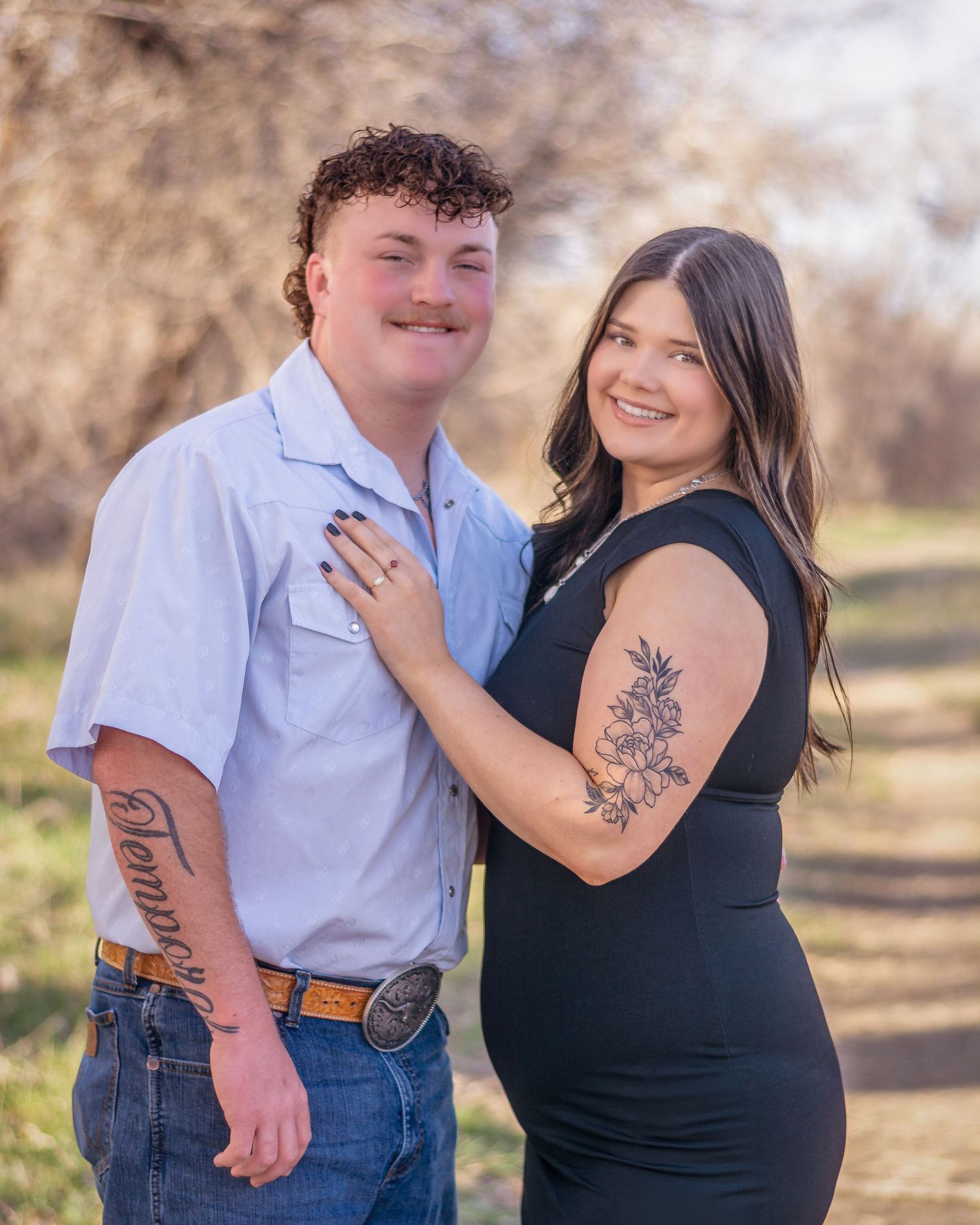 Smiling couple posing outdoors, woman with floral arm tattoo in black dress touching mans chest.