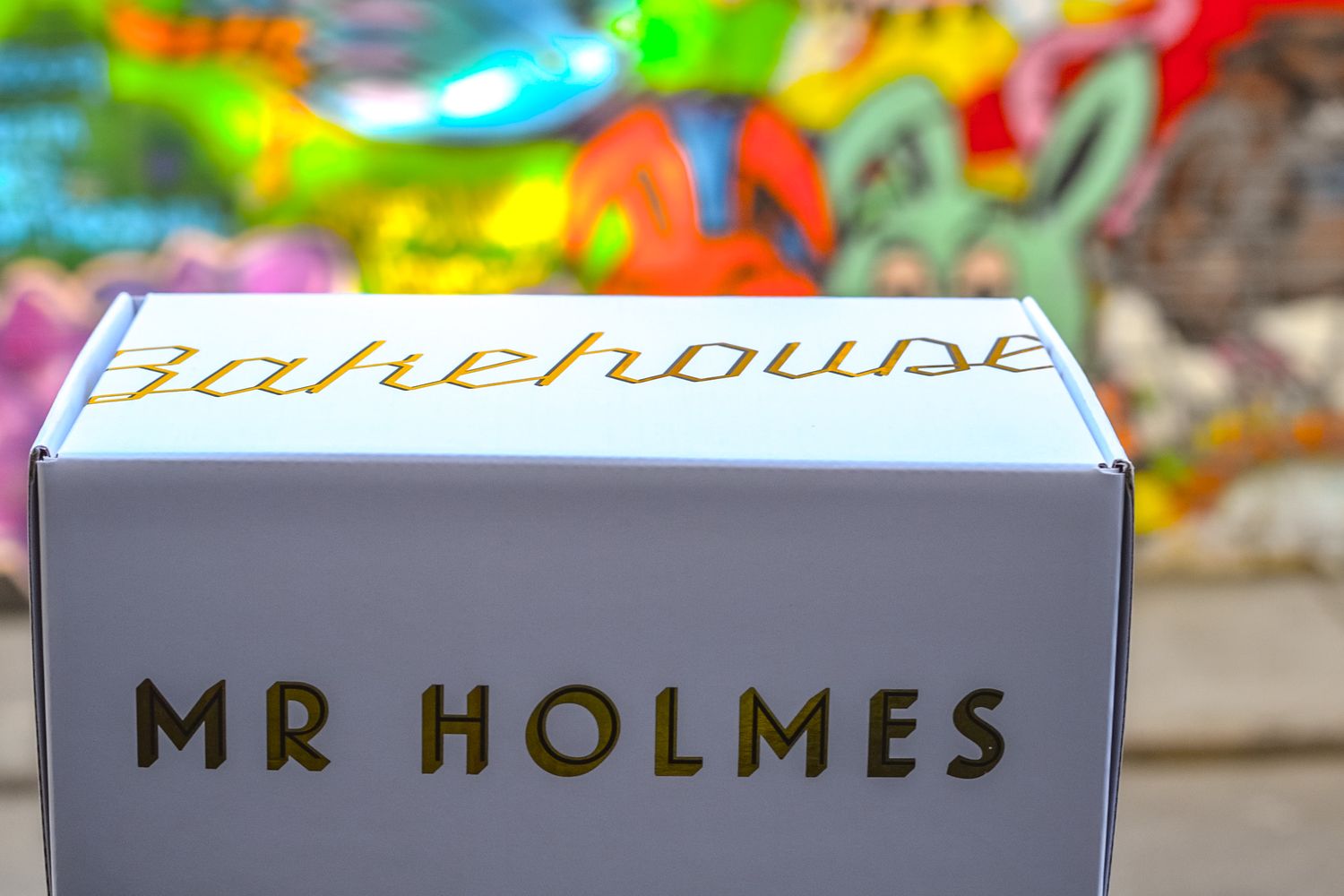 White Mr Holmes Bakehouse pastry box with gold lettering displayed against colorful graffiti wall background.