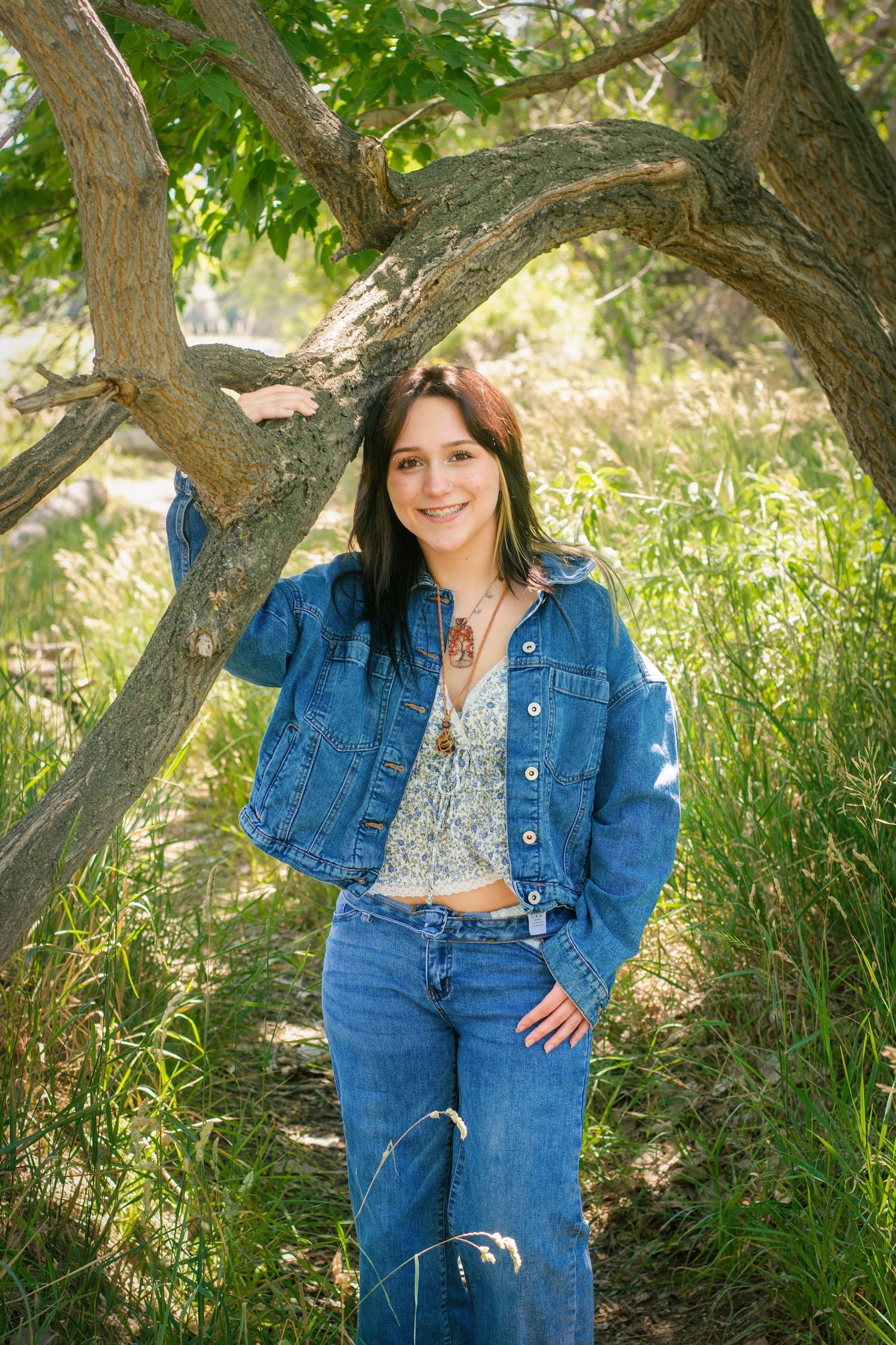 Smiling young woman in denim jacket and jeans posing beside tree in lush green outdoor setting.