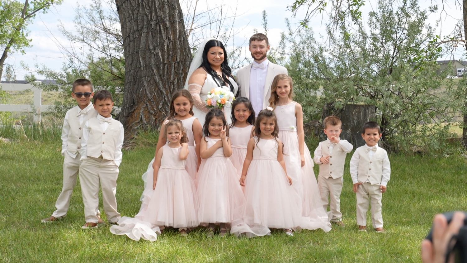 Wedding couple poses with flower girls in pink dresses and ring bearers in white suits outdoors under a tree.
