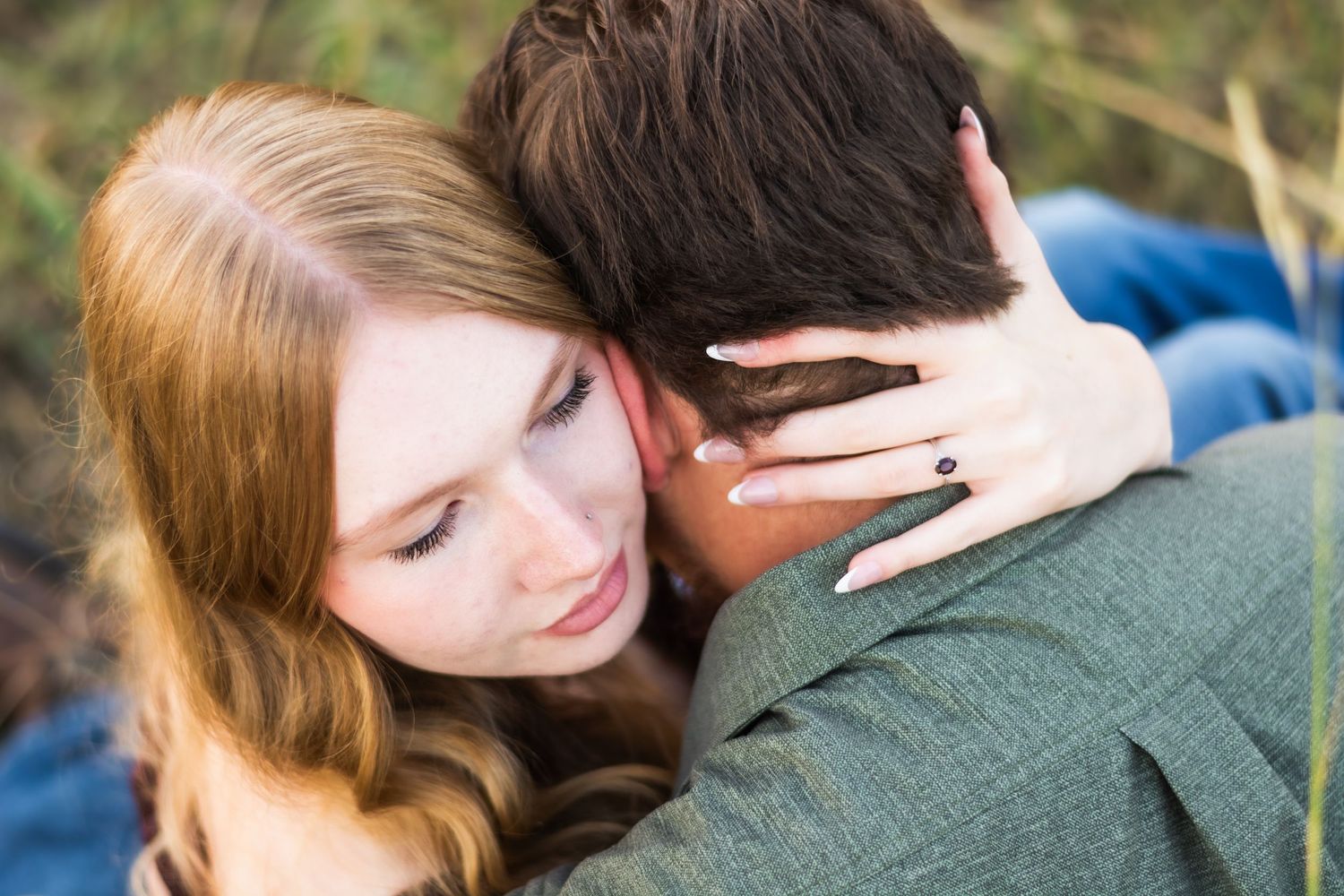 Couple shares a tender embrace outdoors with woman showing engagement ring resting on mans neck.