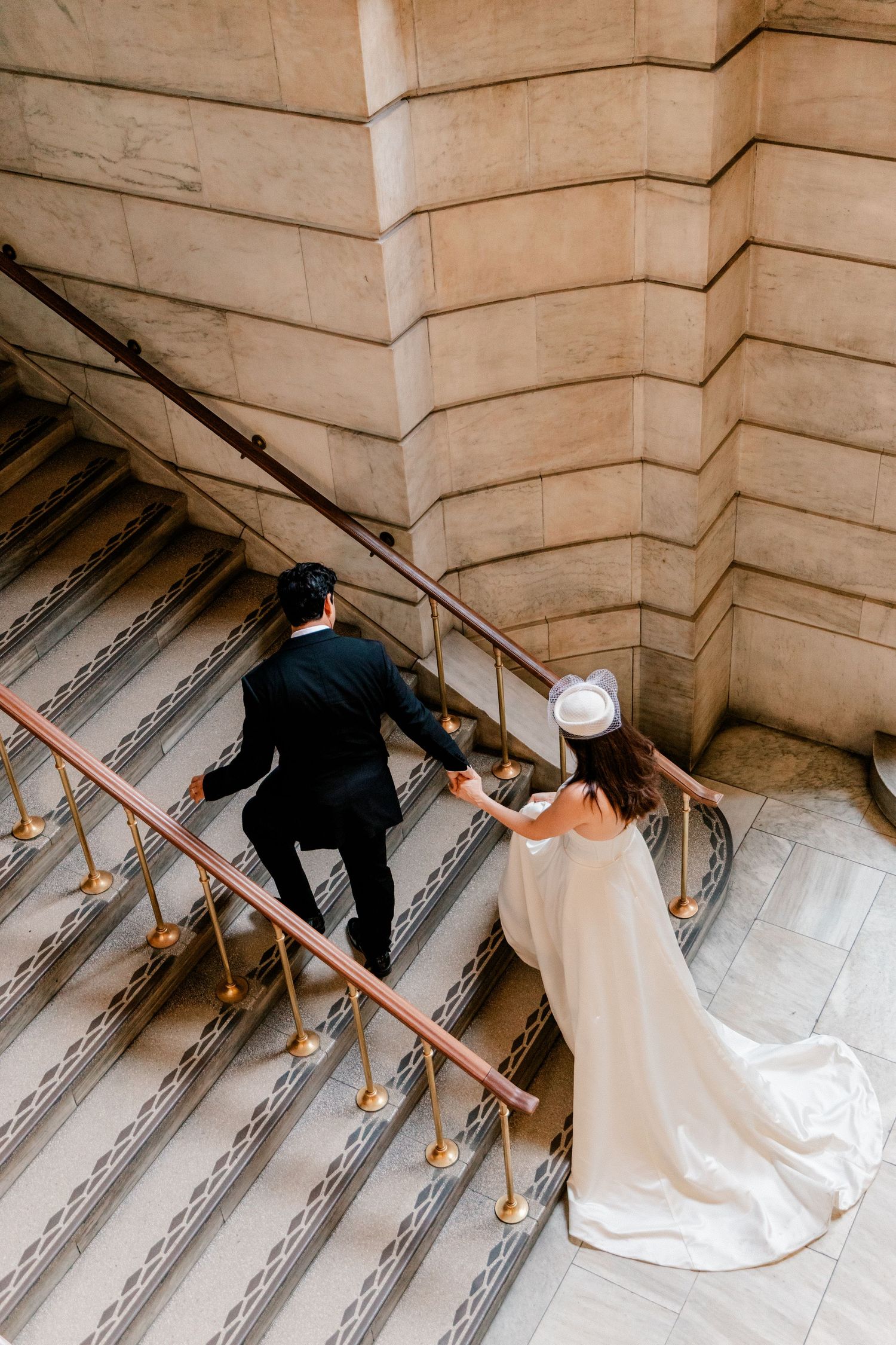 Bride and groom ascending grand stone staircase, bride in white gown with pillbox hat, groom in black suit.