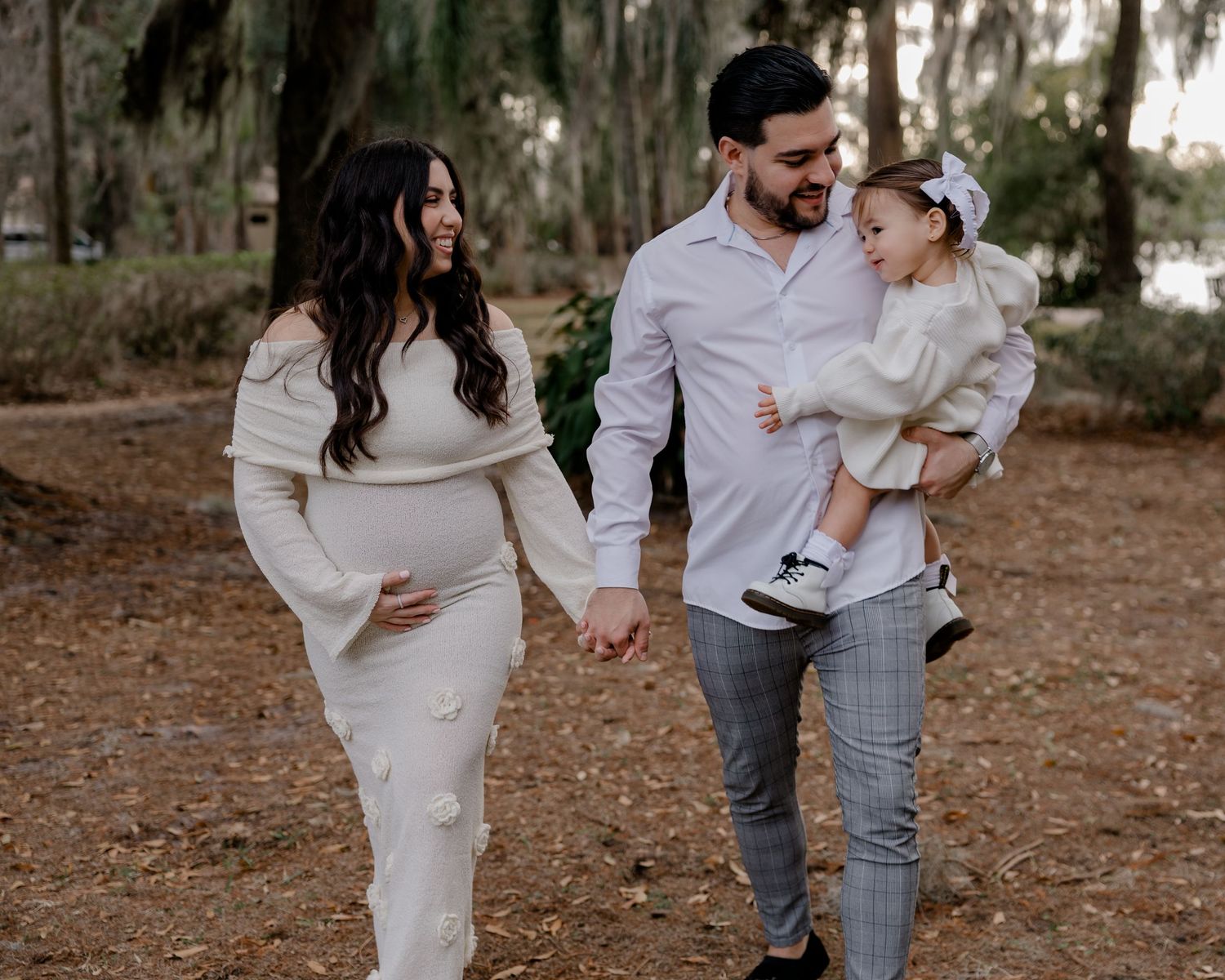 Pregnant woman in white dress walks hand-in-hand with man holding toddler in sunny park setting.