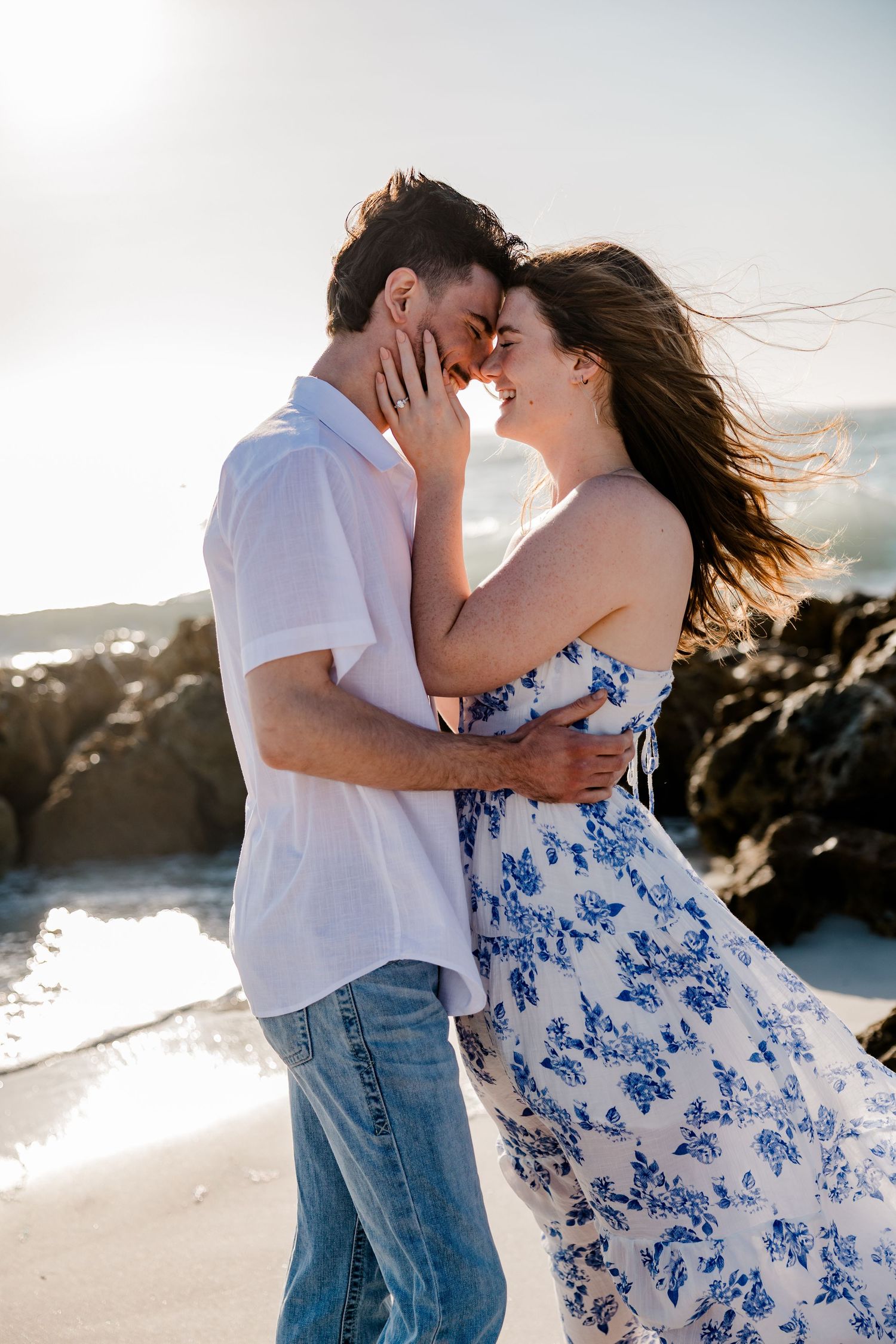 Romantic couple embraces on beach at sunset, woman in flowing blue floral dress and man in white shirt.