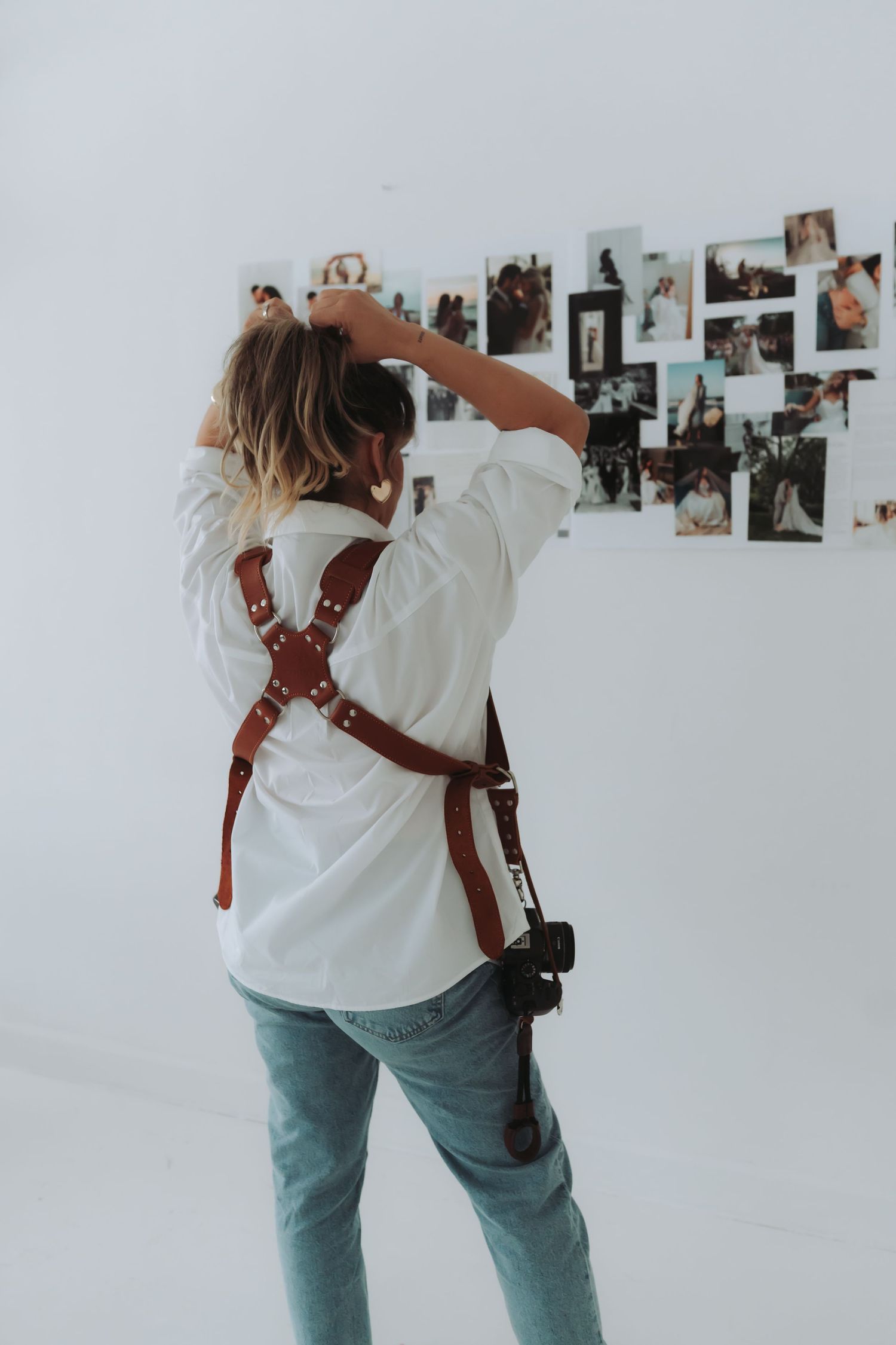Photographer wearing white shirt and leather camera harness stands in front of photo wall with jeans and equipment.