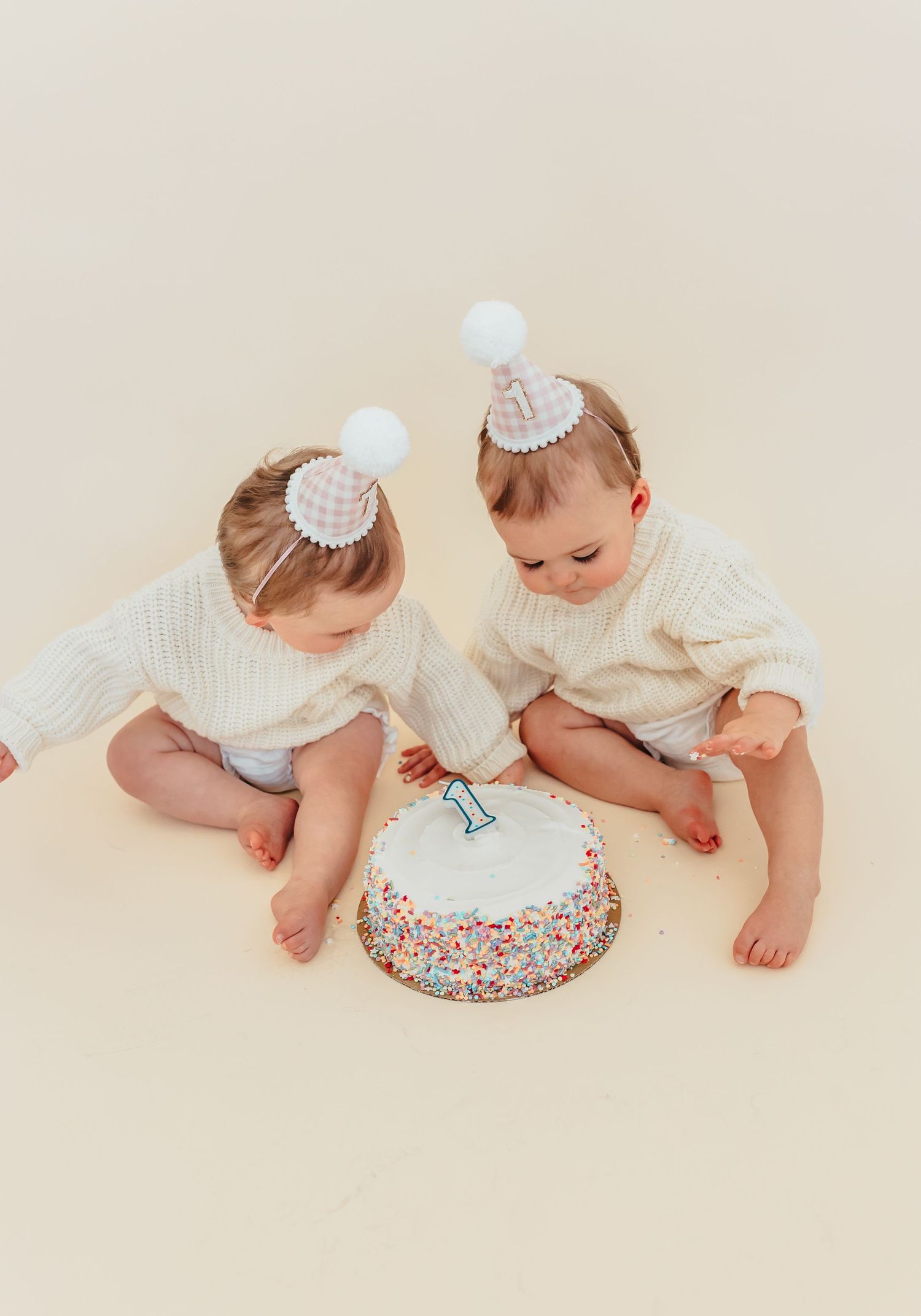 Two infants in white dresses crawl toward a colorful sprinkle-covered cake on a light beige background.