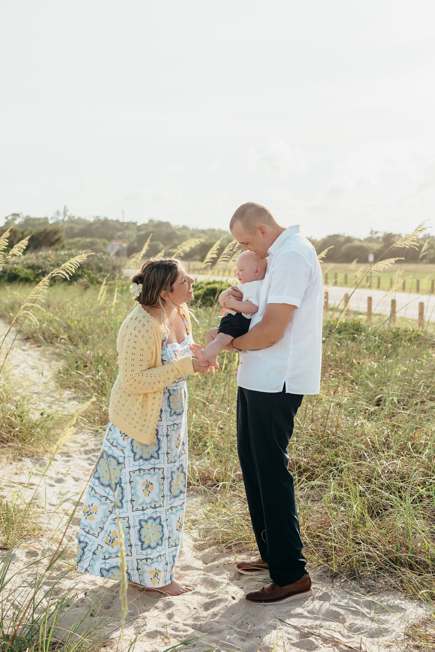 A couple smiles at their newborn baby on a sandy beach path surrounded by sea oats on a sunny day.