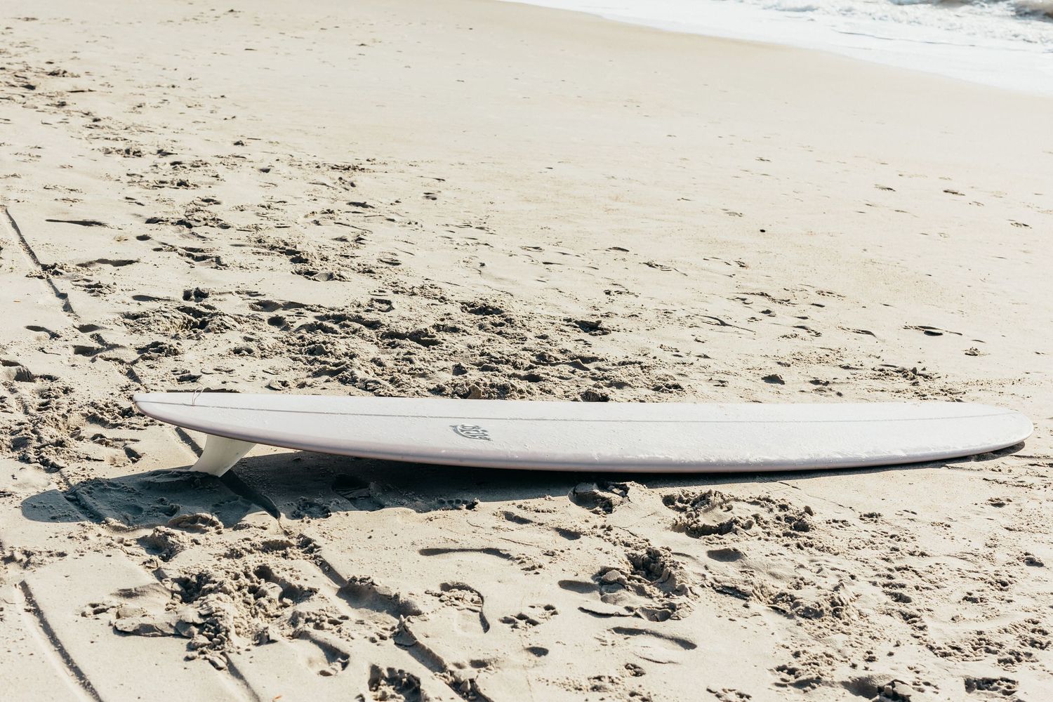 White surfboard resting on sandy beach near ocean waves on a sunny day.