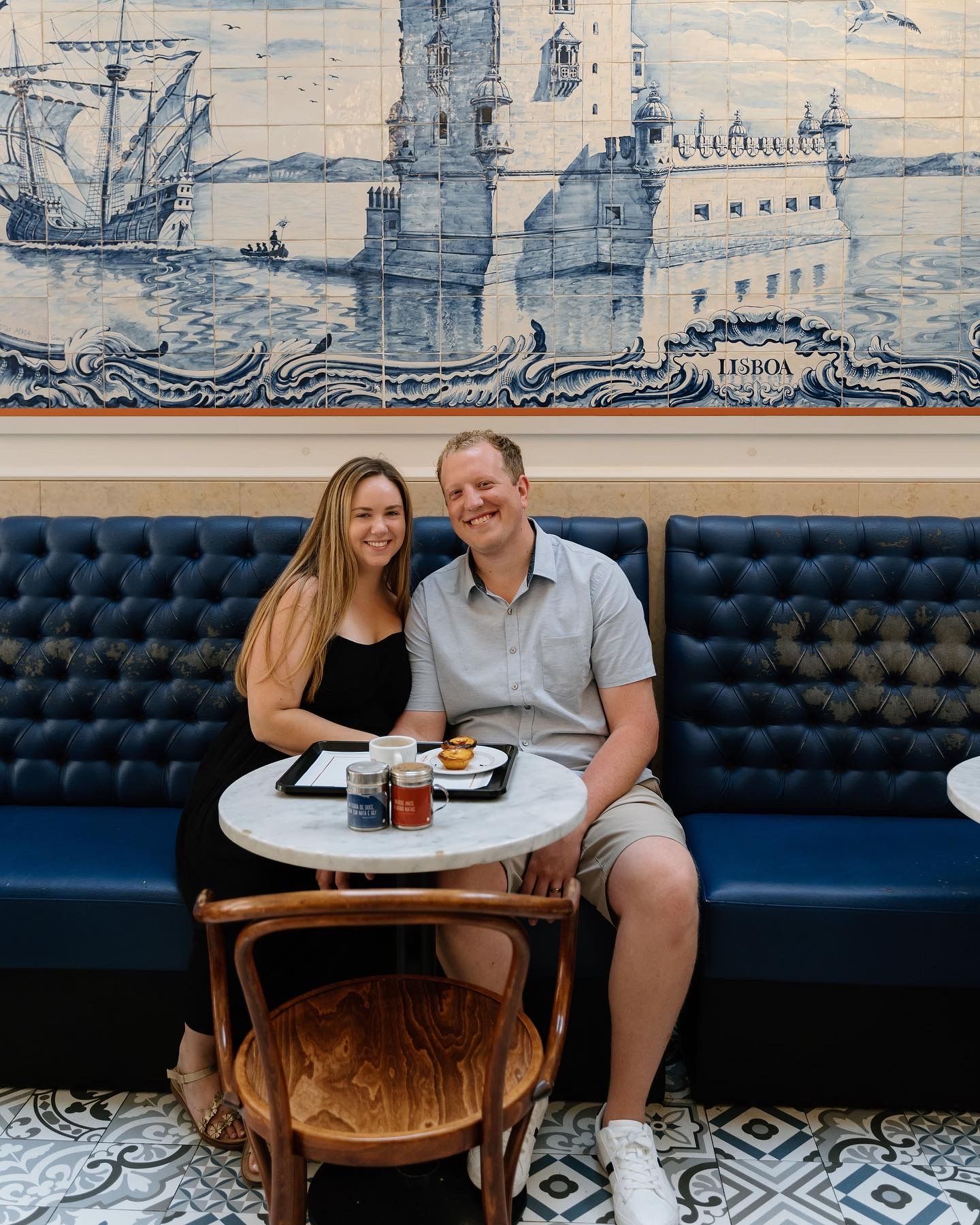 A couple sits at a restaurant booth with blue tufted seating and decorative tile flooring enjoying a meal together.