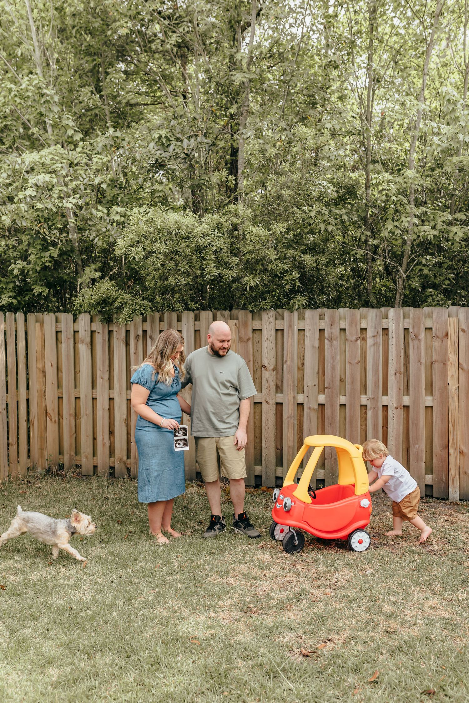 Family with toddler and dog in backyard, holding ultrasound photo by wooden fence with Little Tikes car.