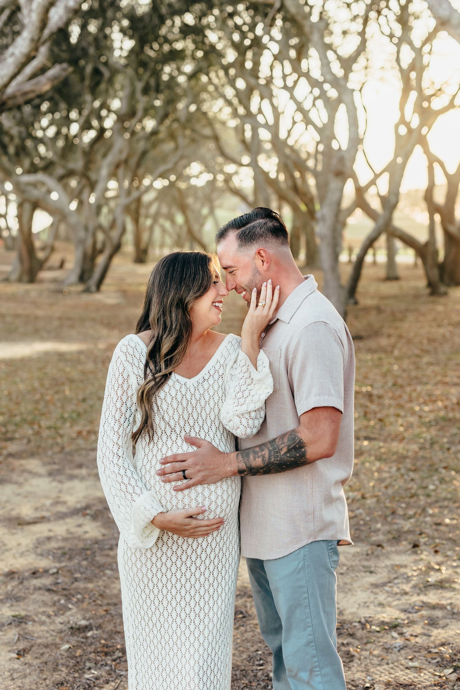A couple shares a tender moment outdoors, the woman in a white knit maternity dress cradling her bump as they touch foreheads.