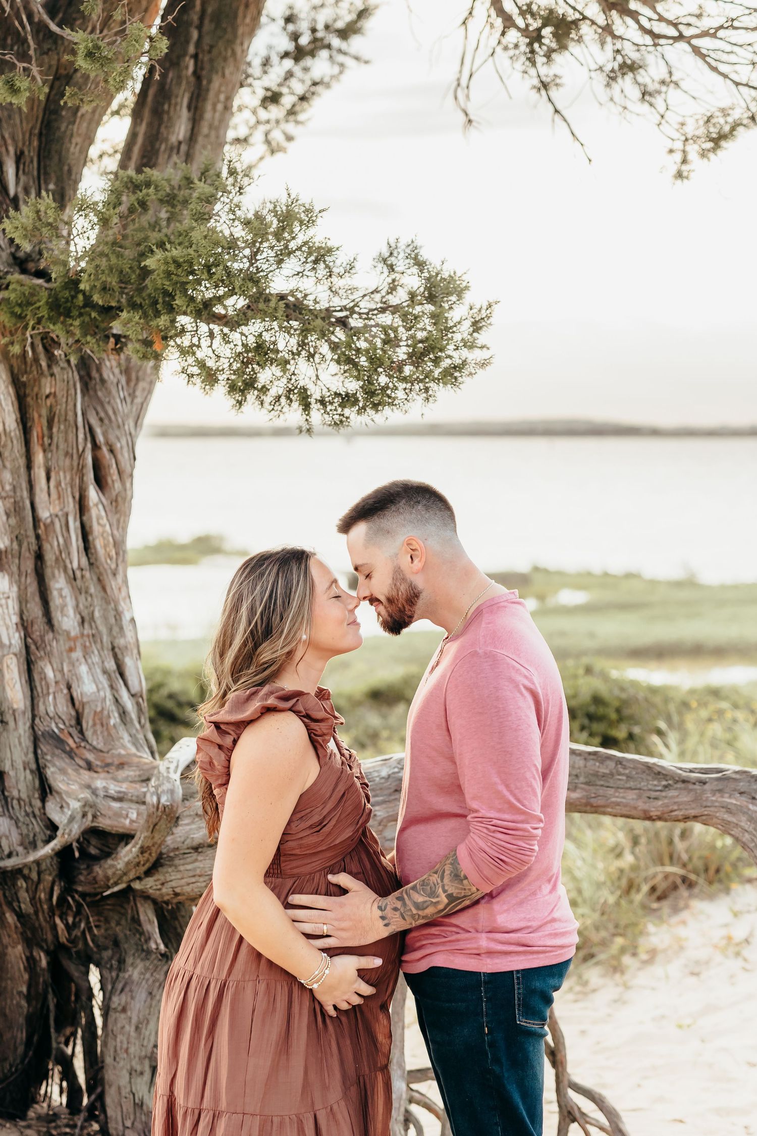 A couple in matching pink and brown outfits share intimate moments during a maternity photoshoot on a beach.