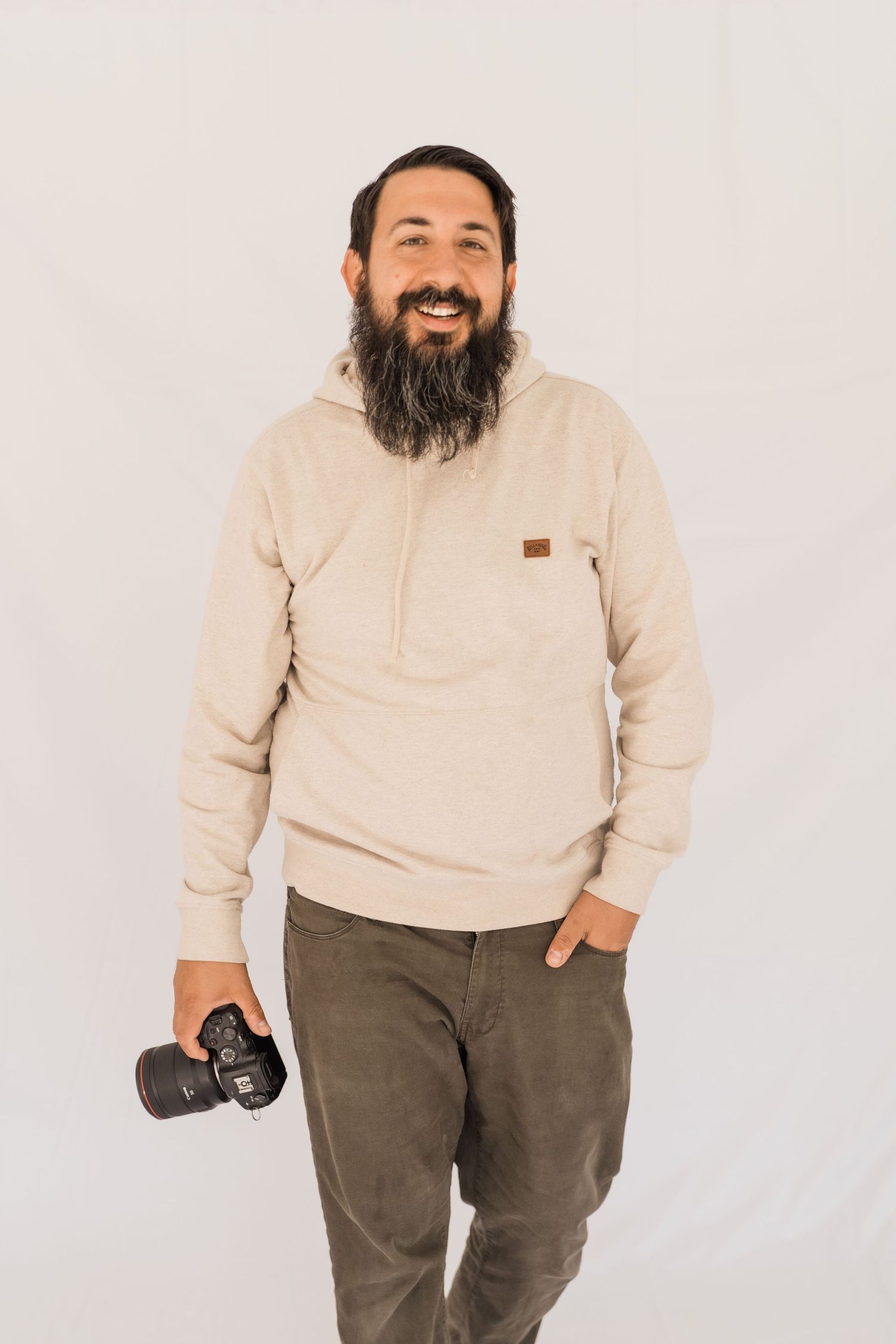Photographer in beige sweater and brown pants holds professional camera against white background.