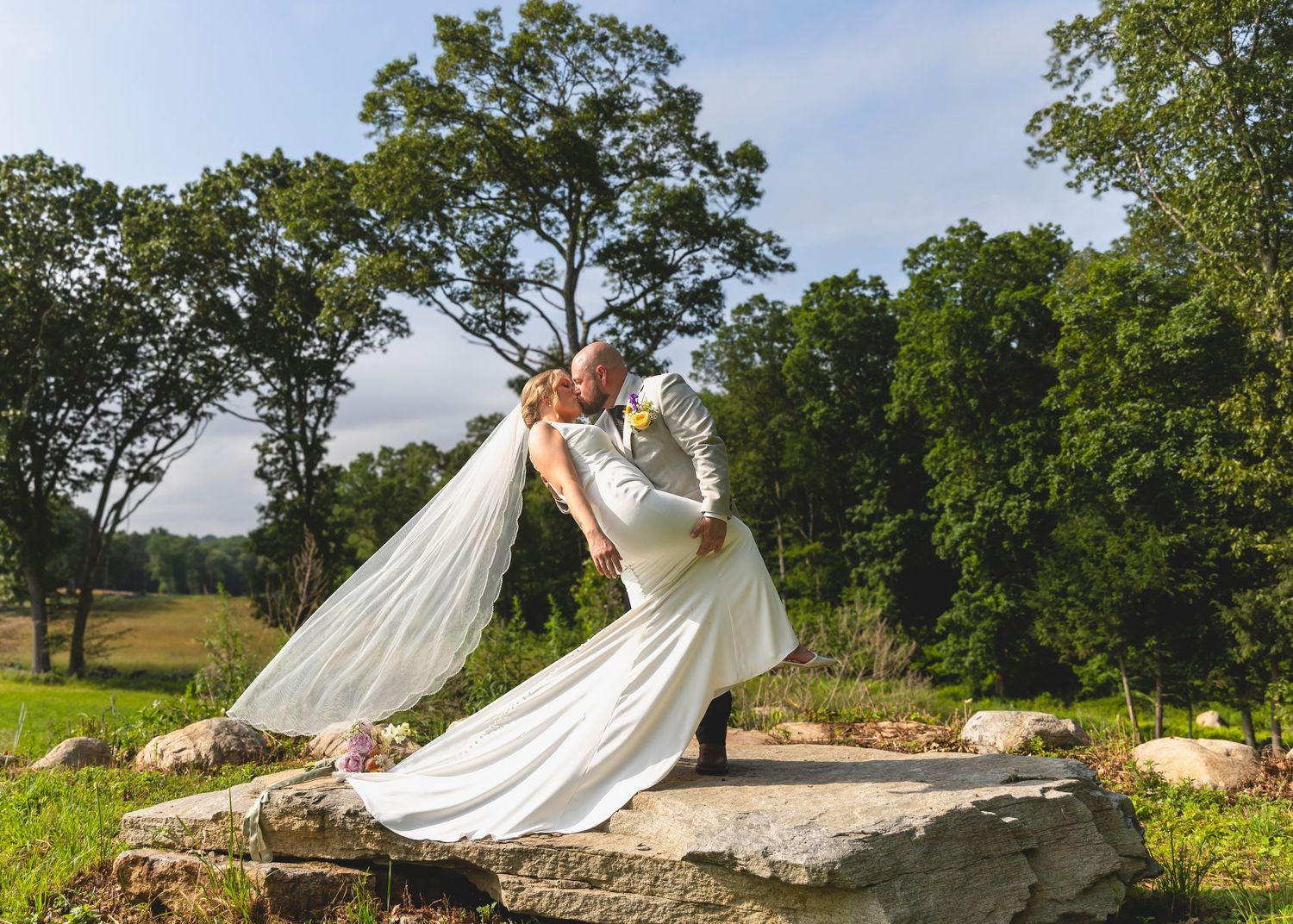 A romantic couple in white formal attire share a dramatic dip pose on a stone wall surrounded by lush green trees.