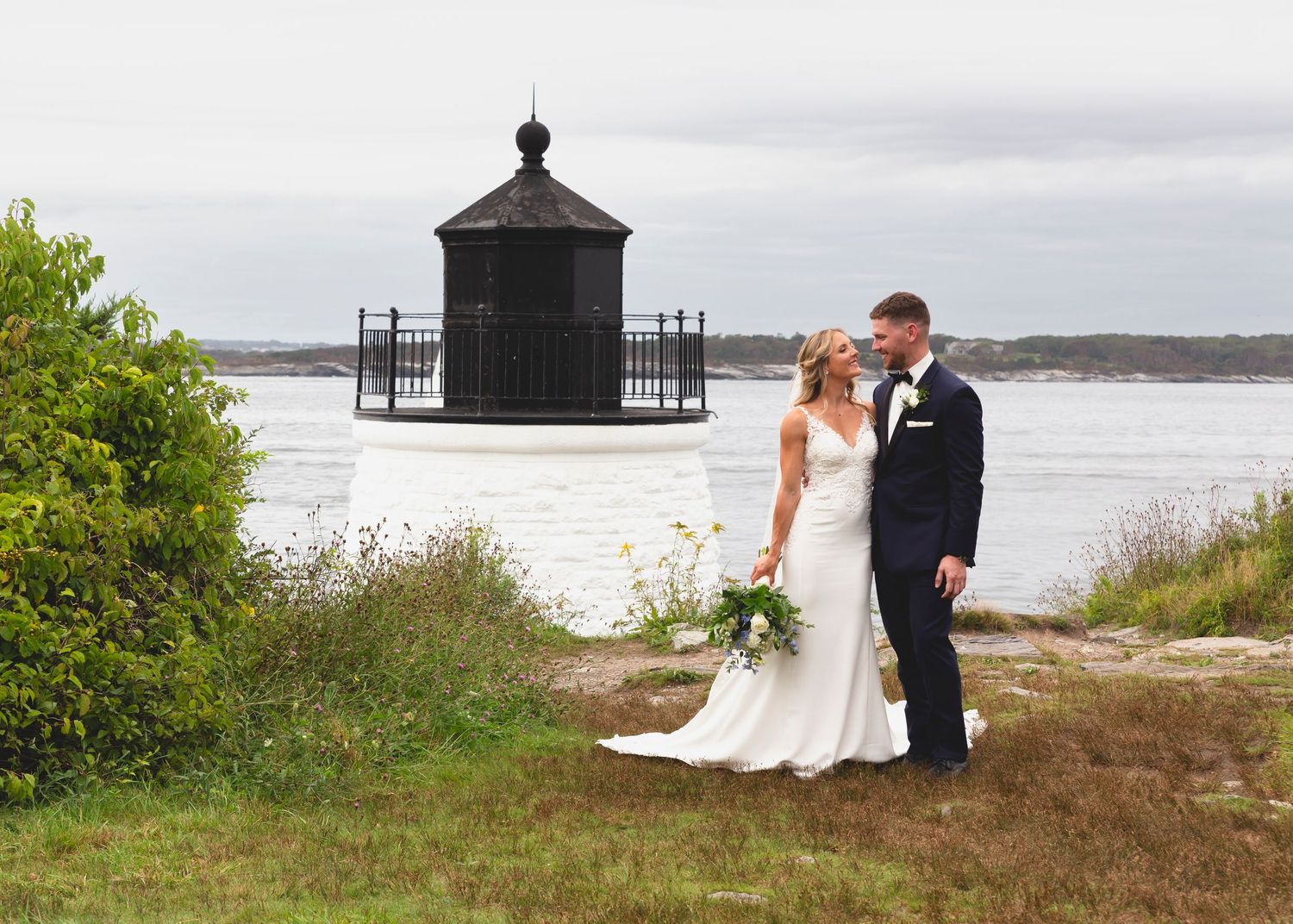 A wedding couple poses by a historic coastal lighthouse structure with water and greenery in the scenic backdrop.