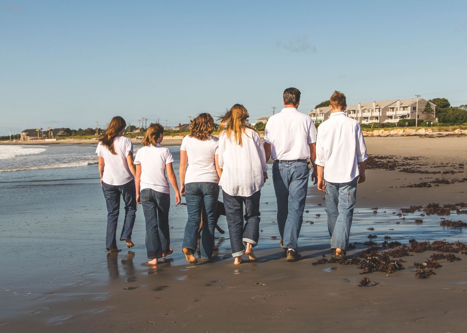 Group in matching white shirts and jeans walking together on a beach at sunset with coastal buildings in background.