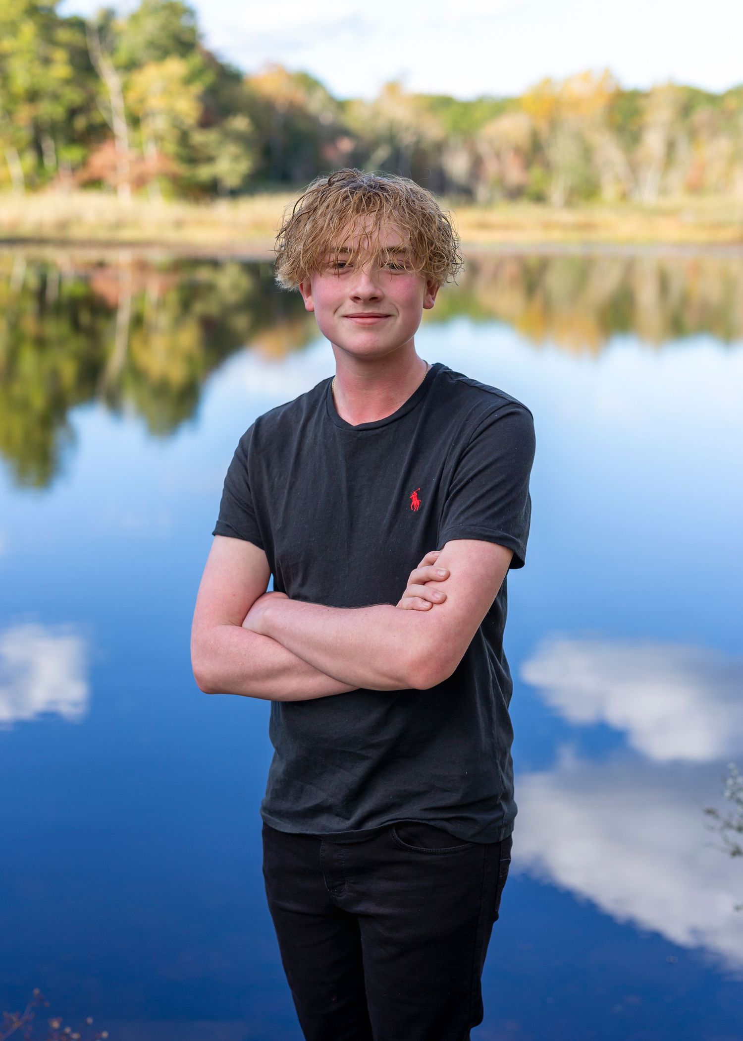 Senior photo of teen in black jeans and black shirt with water and trees in the background