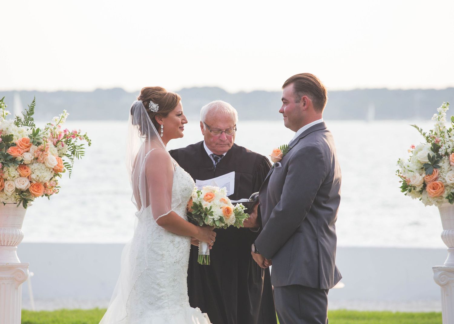 A scenic waterfront wedding ceremony with an officiant and couple exchanging vows beside floral arrangements.