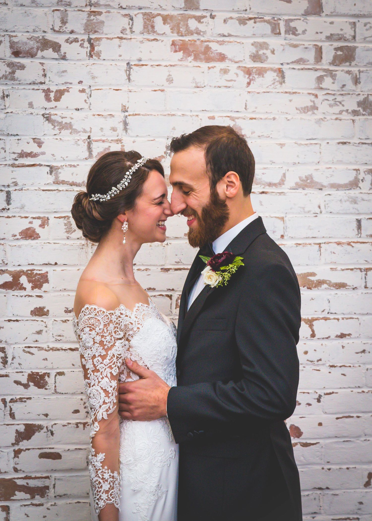 A romantic wedding moment with a couple in white dress and black suit embracing against a weathered brick wall.