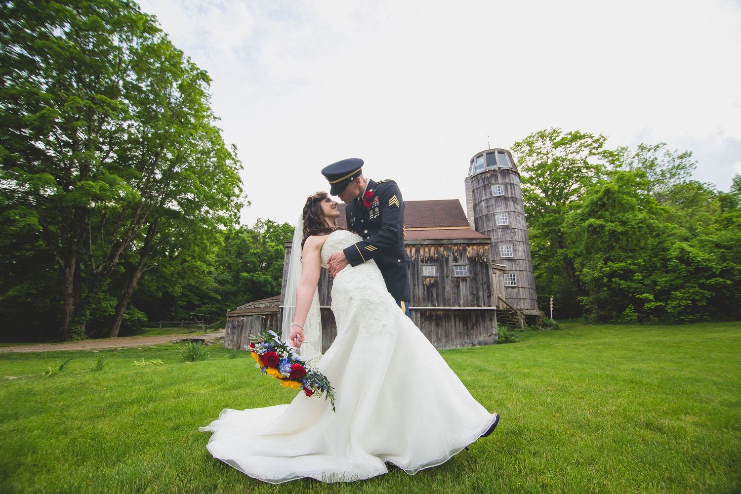 Wedding couple dances on lawn with historic farm building in background.