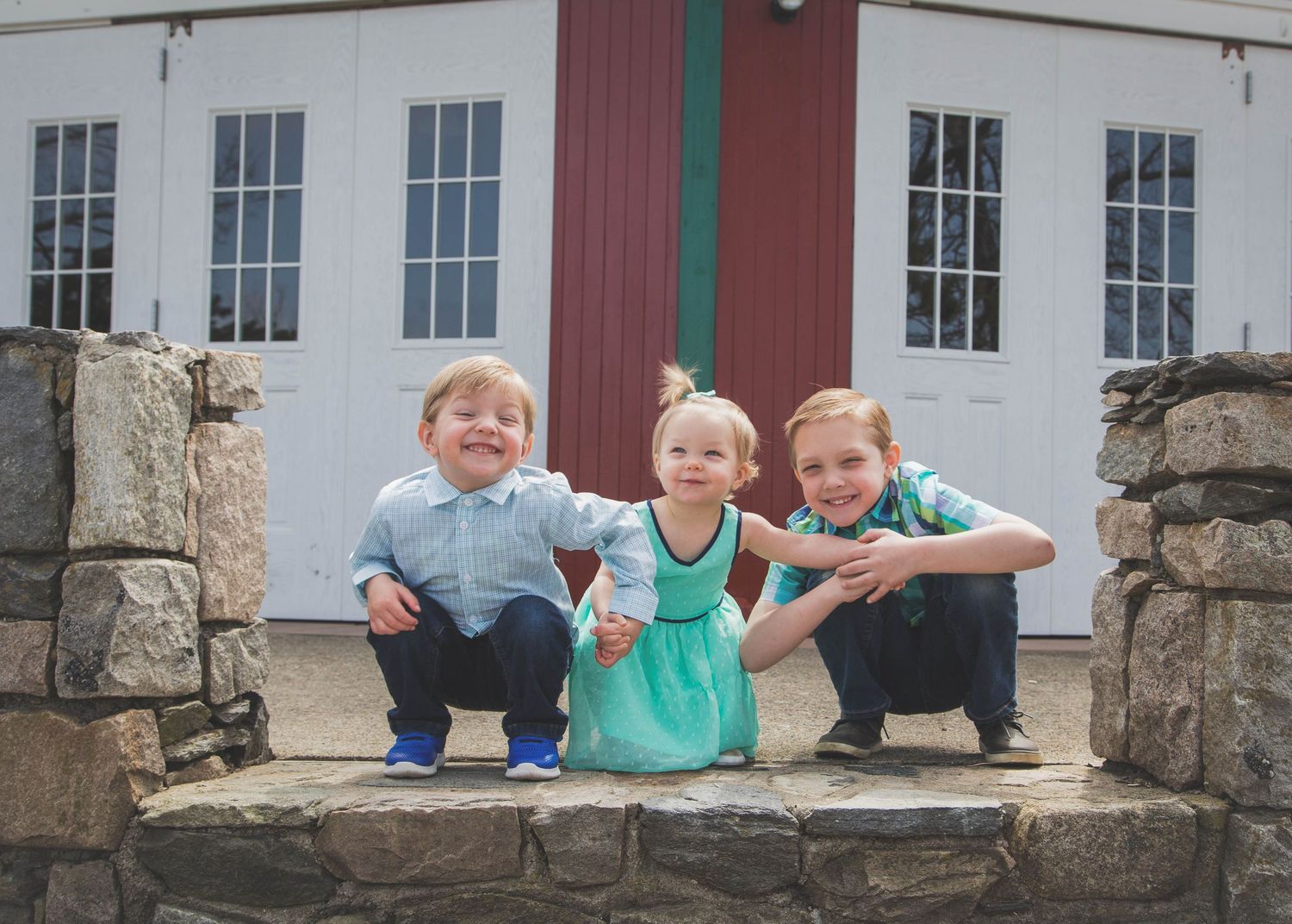 Three young children sit together on stone steps in front of a white building with windows.