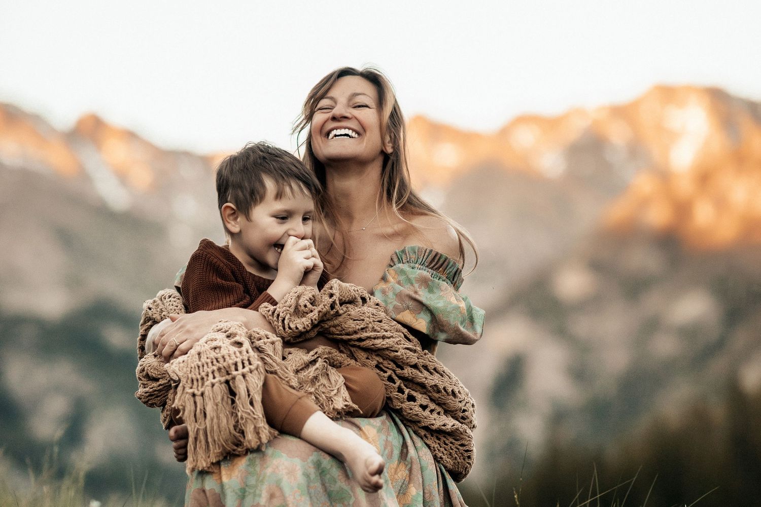 A joyful mother holds her young son in a mountain meadow at golden hour, both wrapped in cozy earth-toned clothing.