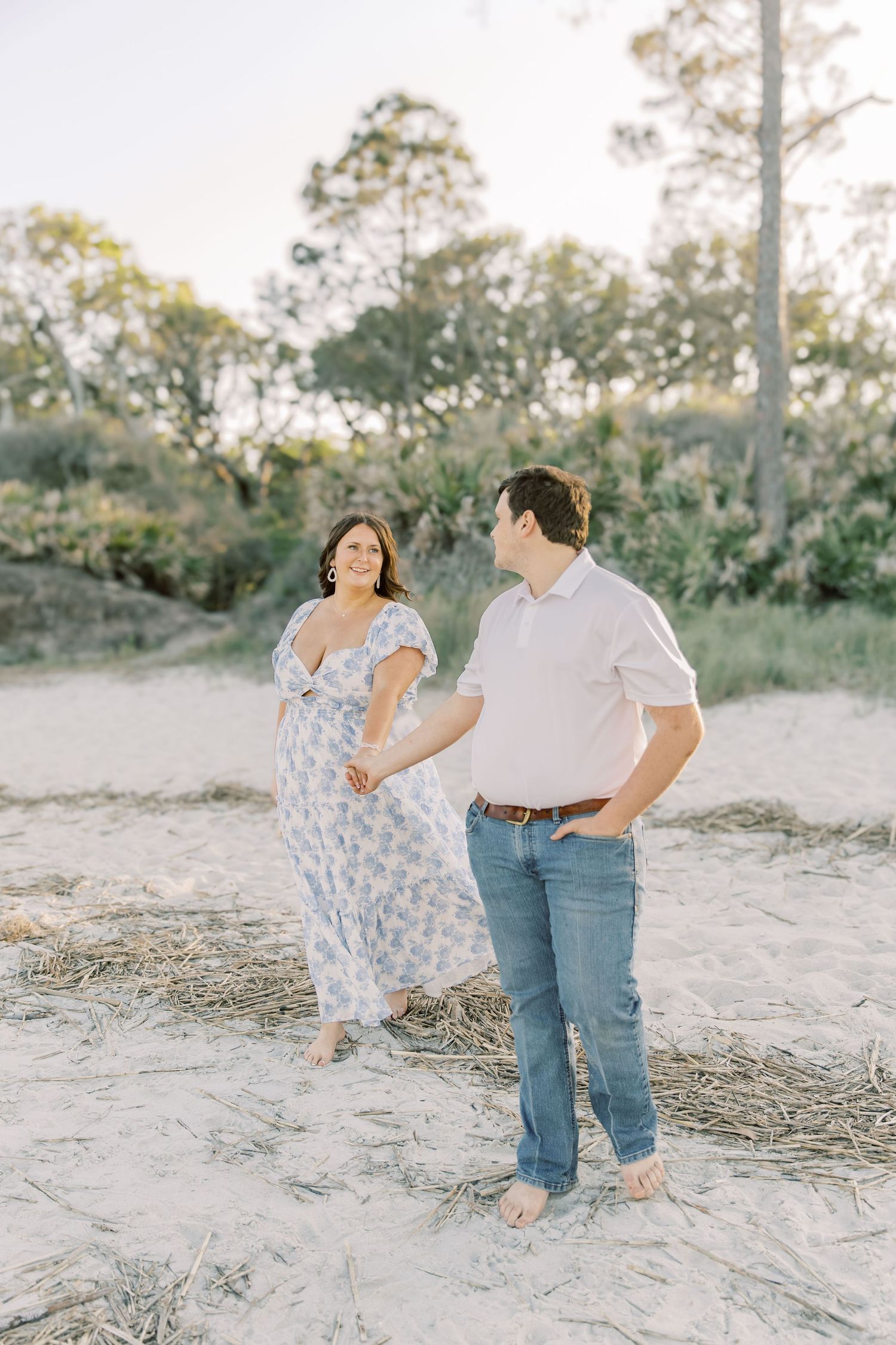 A couple holds hands while walking barefoot on a sandy beach path surrounded by trees in soft evening light.