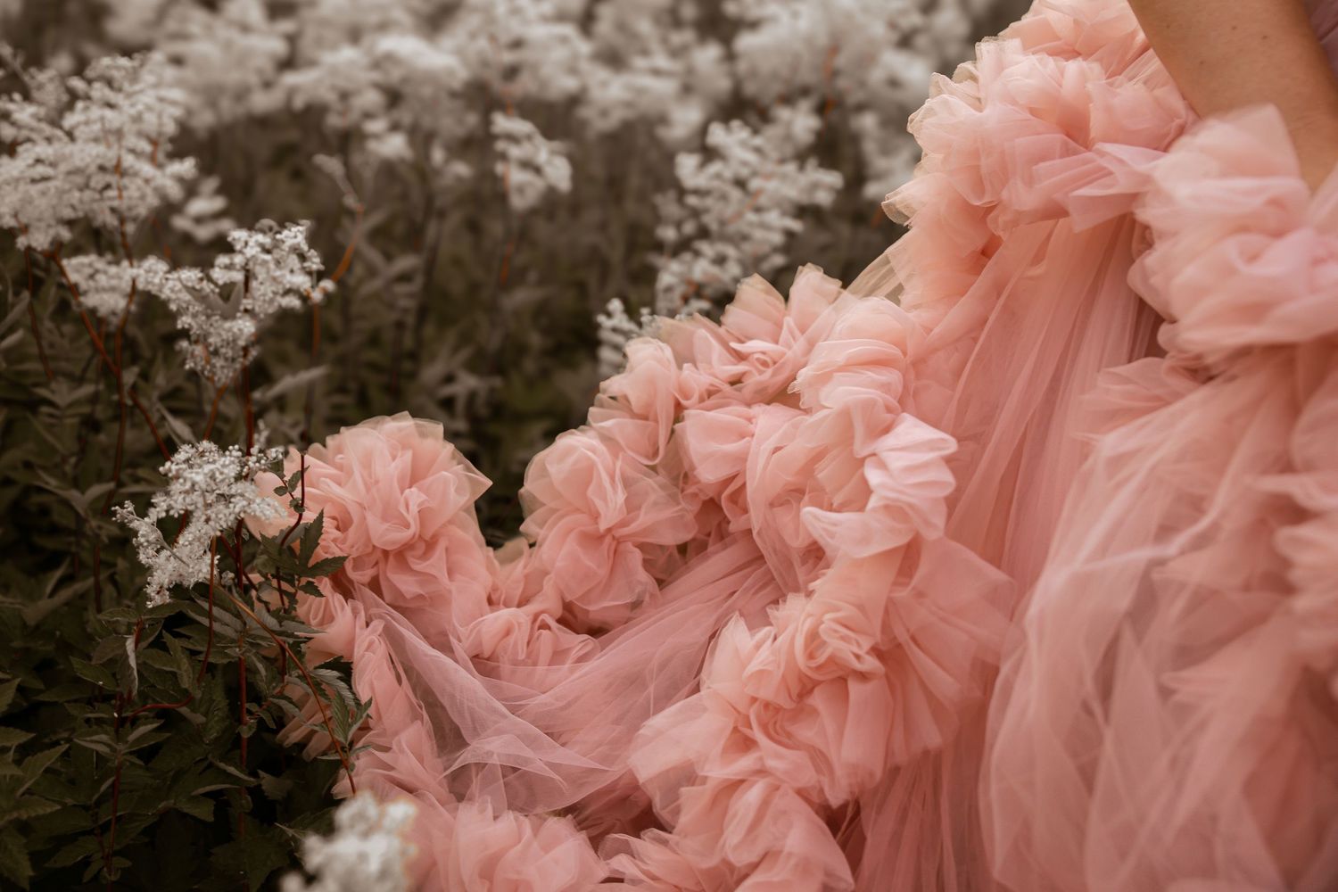 A billowing pink tulle gown with ruffled details flows gracefully among white wildflowers in a dreamy outdoor setting.