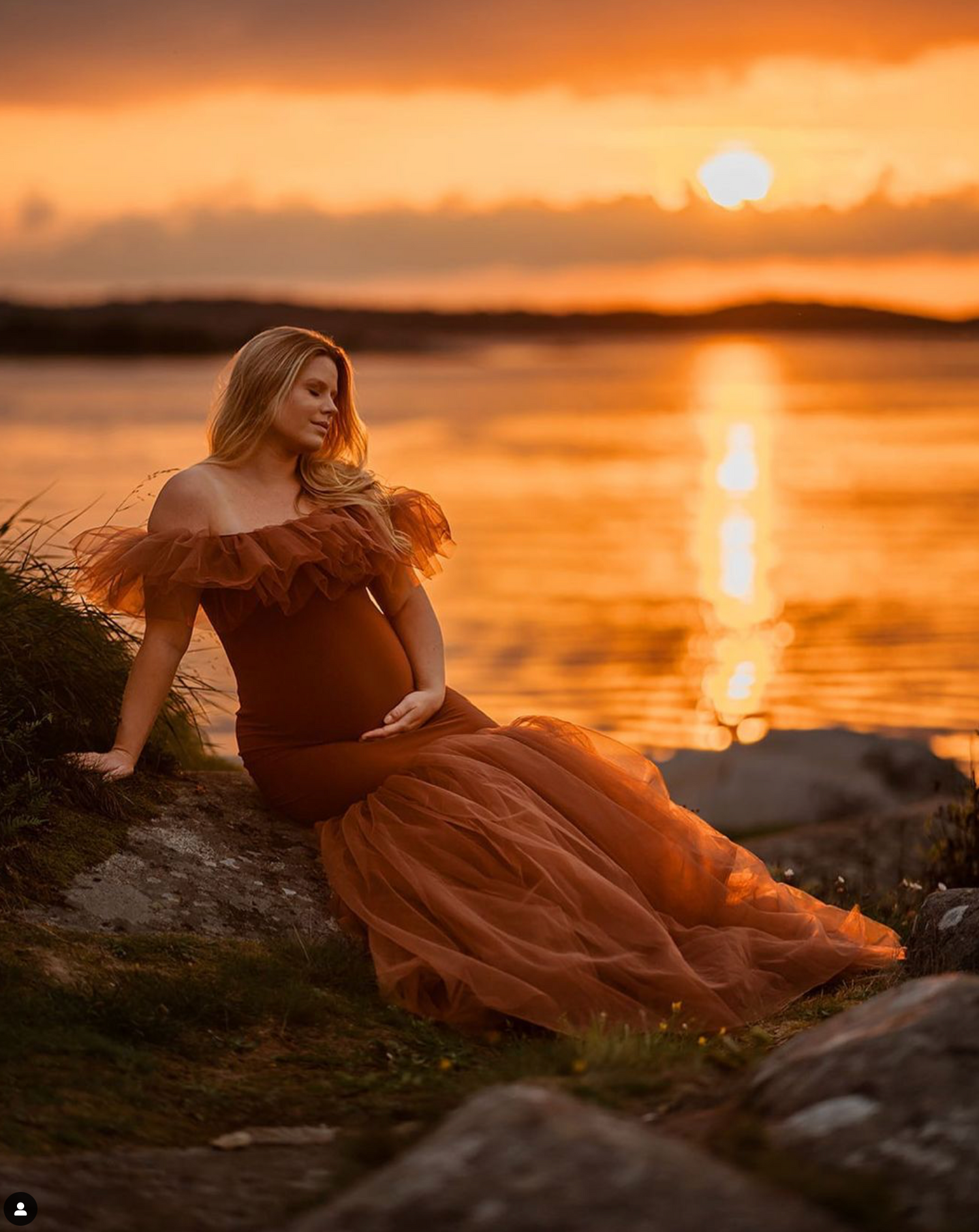 A flowing brown dress catches golden light as a figure sits by the water during a stunning coastal sunset.