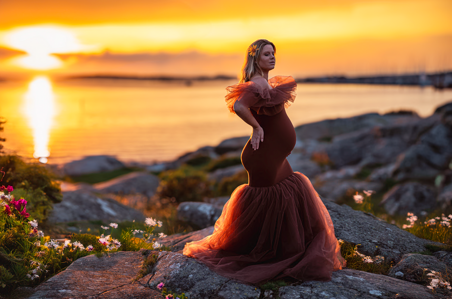 A silhouetted figure in a flowing red gown poses on coastal rocks at sunset with golden light reflecting on calm waters.