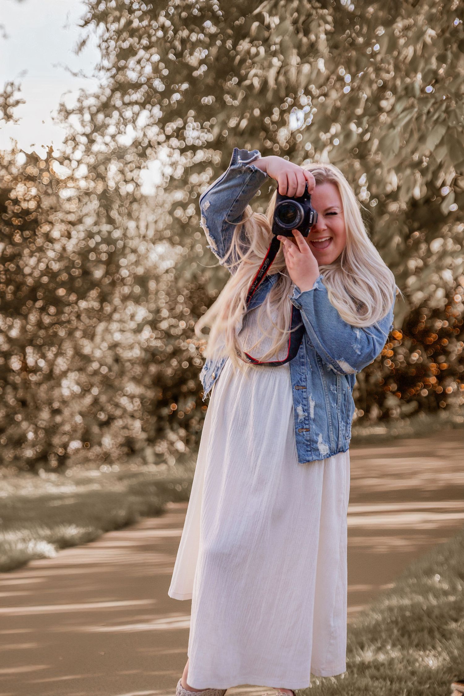 Photographer in white maxi dress and denim jacket holds camera in outdoor garden setting during golden hour.