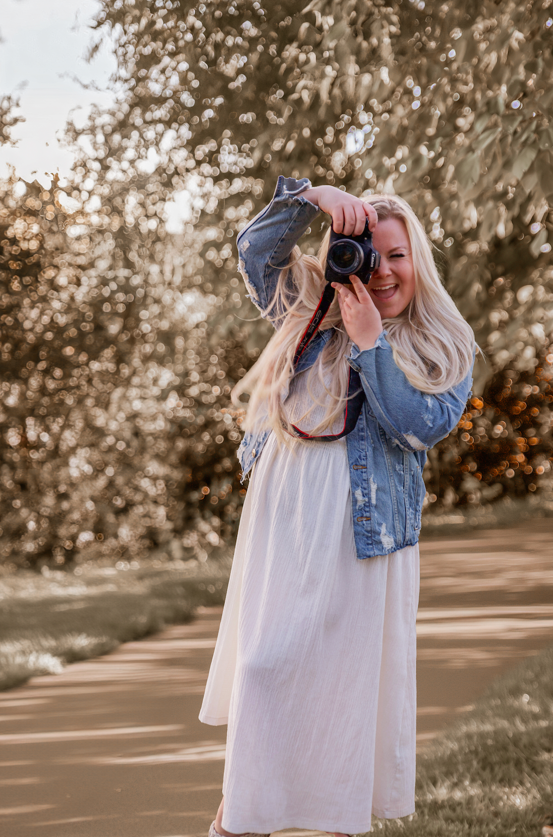 Photographer in white dress and denim jacket holds professional camera outdoors during golden hour.