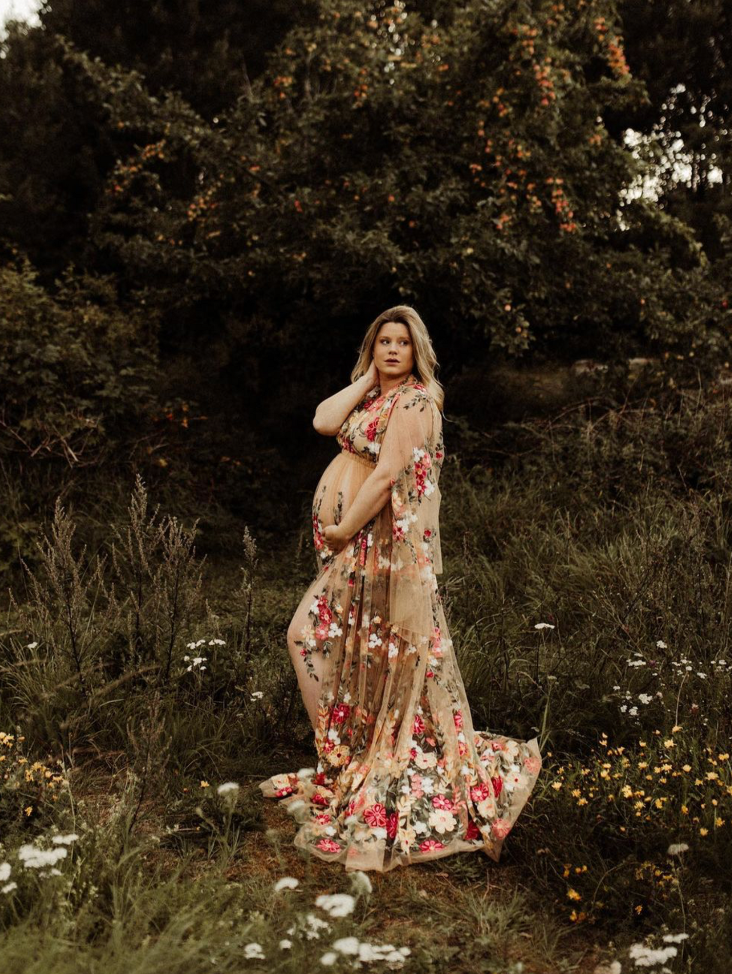 Expecting mother wearing a floral maxi dress poses gracefully in a wildflower meadow at sunset.