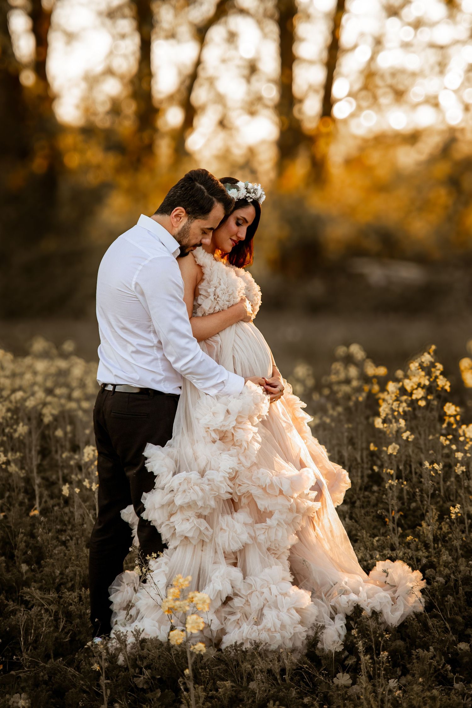 A couple in formal attire share an intimate moment at sunset in a wildflower field, her ruffled dress catching golden light.