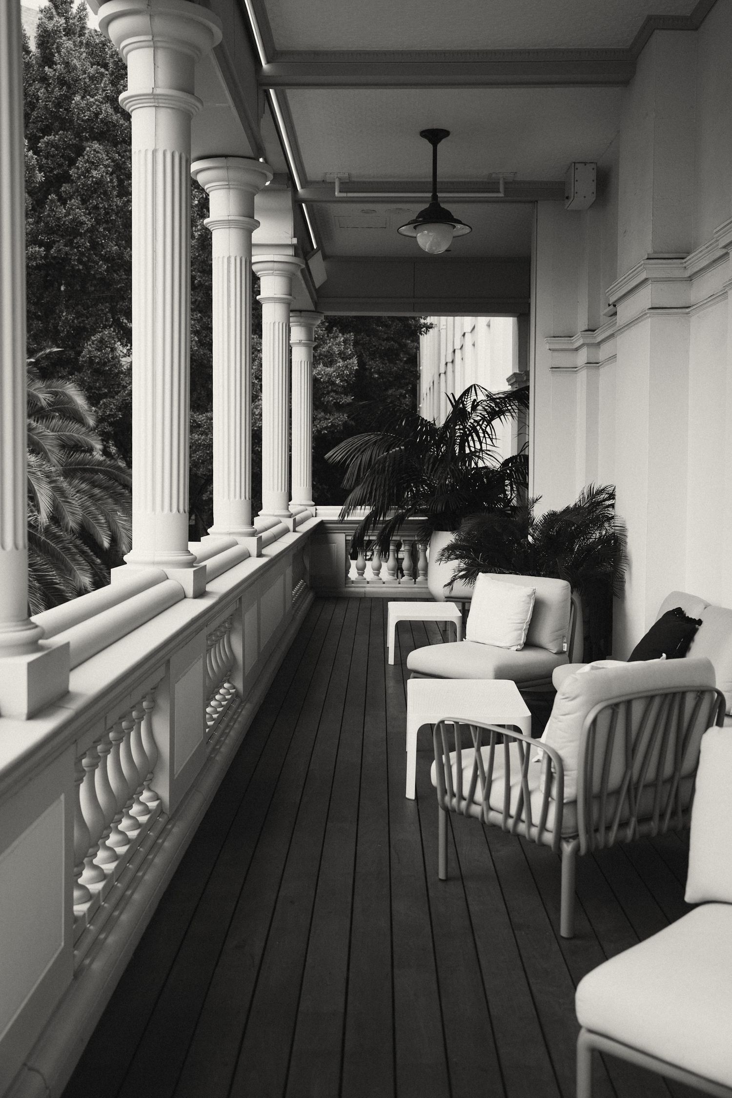 Black and white view of an elegant porch with white columns, wicker furniture, and dark wooden decking.
