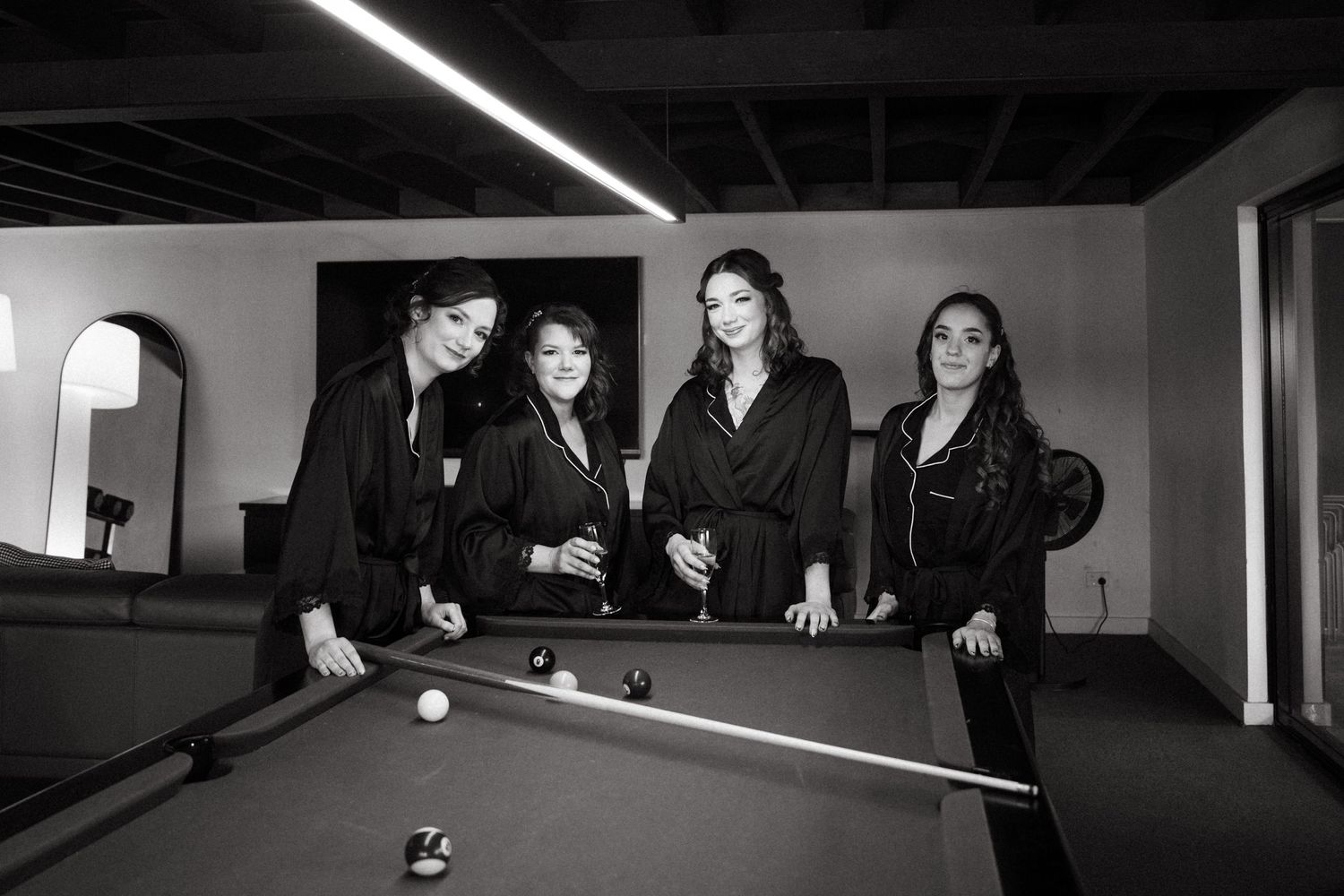 Four women in black attire pose around a pool table in a stylish monochrome photo shoot setting.
