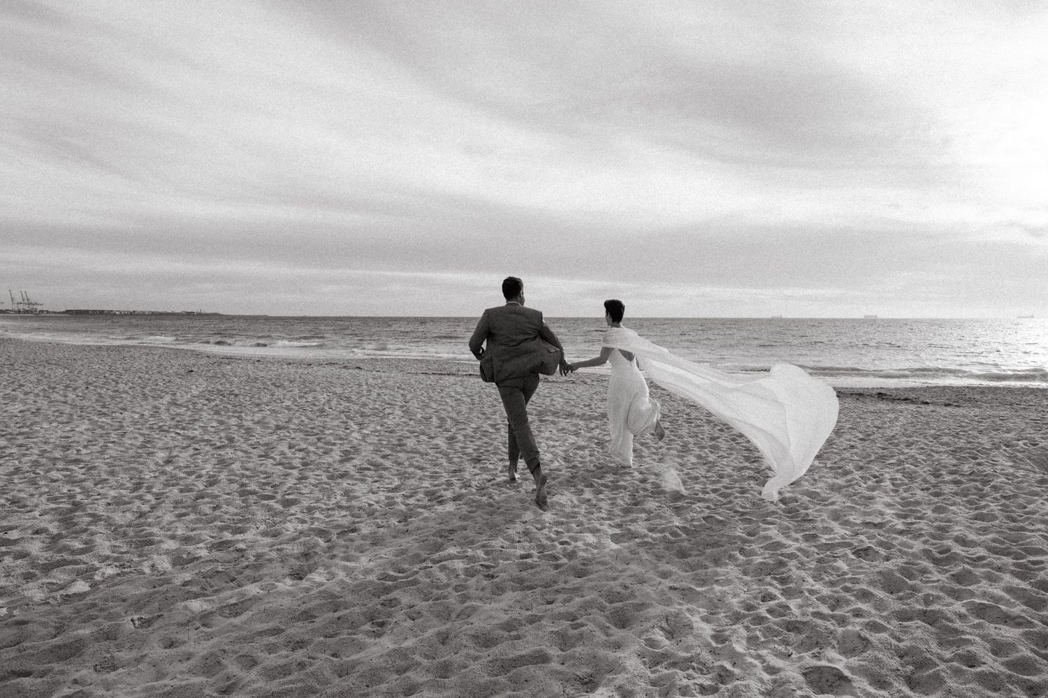 Black and white photo of a bride and groom walking hand in hand on a sandy beach, her veil flowing in the wind.