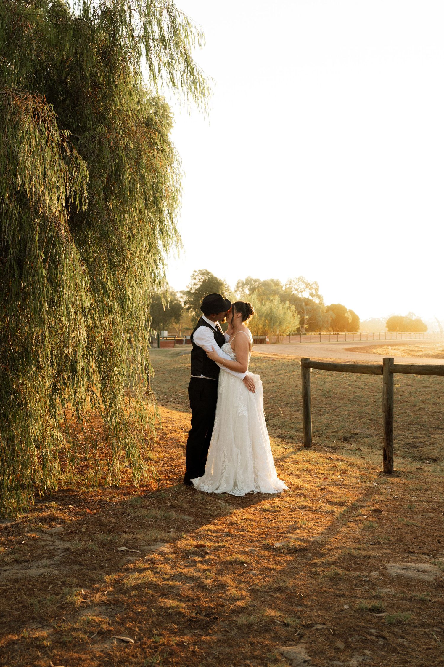 Bride and groom share a romantic kiss under a weeping willow tree at golden hour on a rustic farm wedding venue.