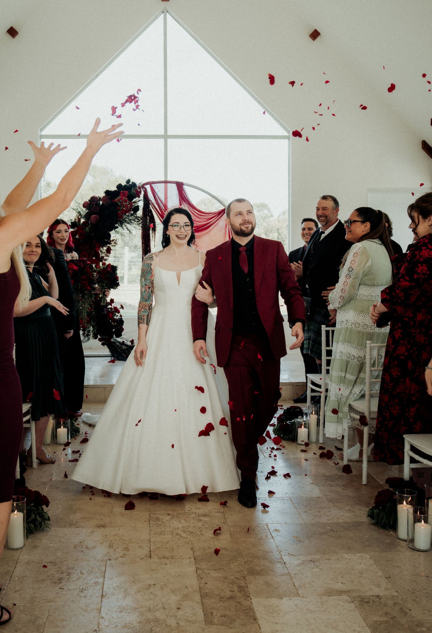Newlyweds walk down the aisle as guests toss red rose petals during an elegant indoor wedding ceremony.