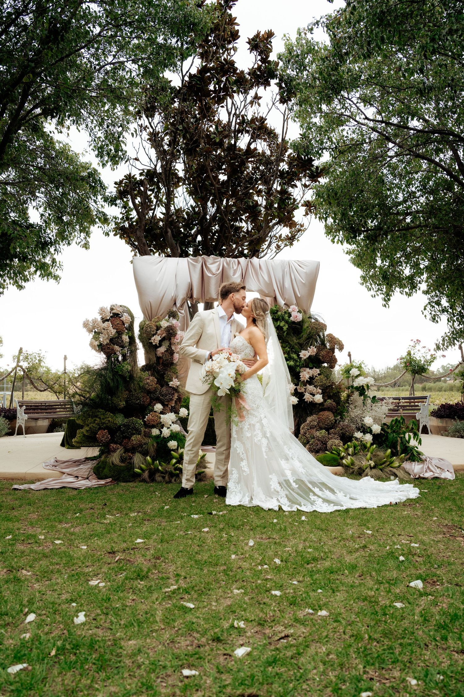 Bride and groom kissing at floral wedding altar with hydrangeas and greenery under trees.