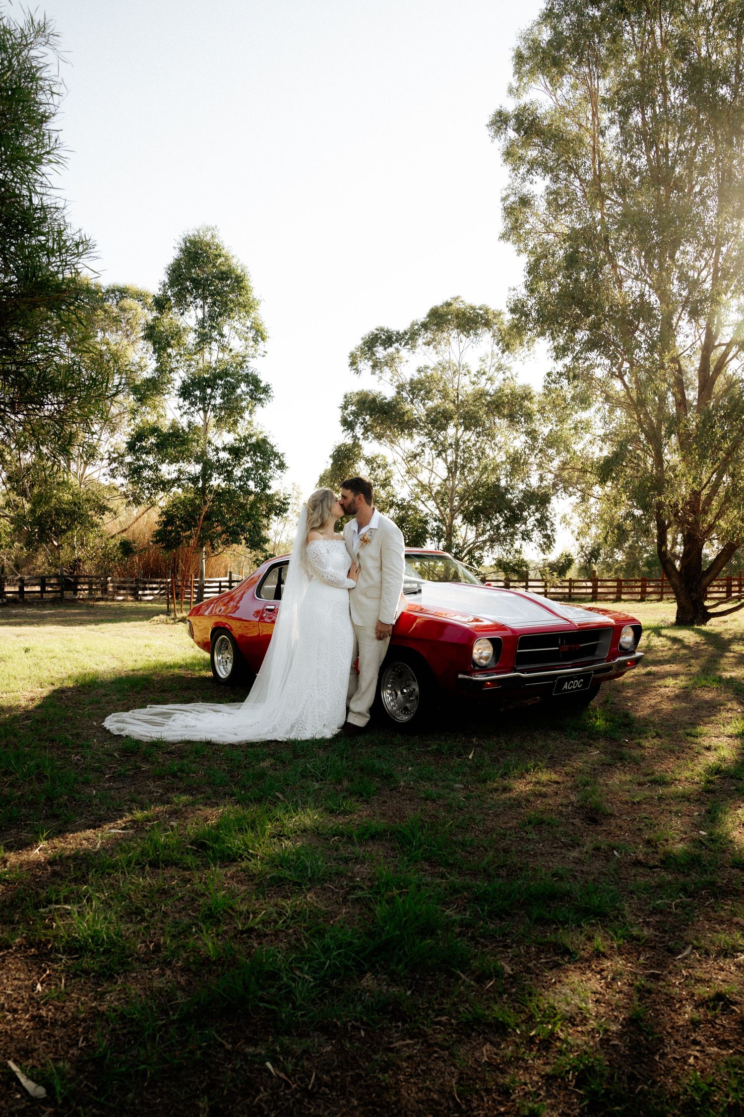 Bride and groom kissing beside a classic red muscle car on a sunny green lawn with eucalyptus trees.