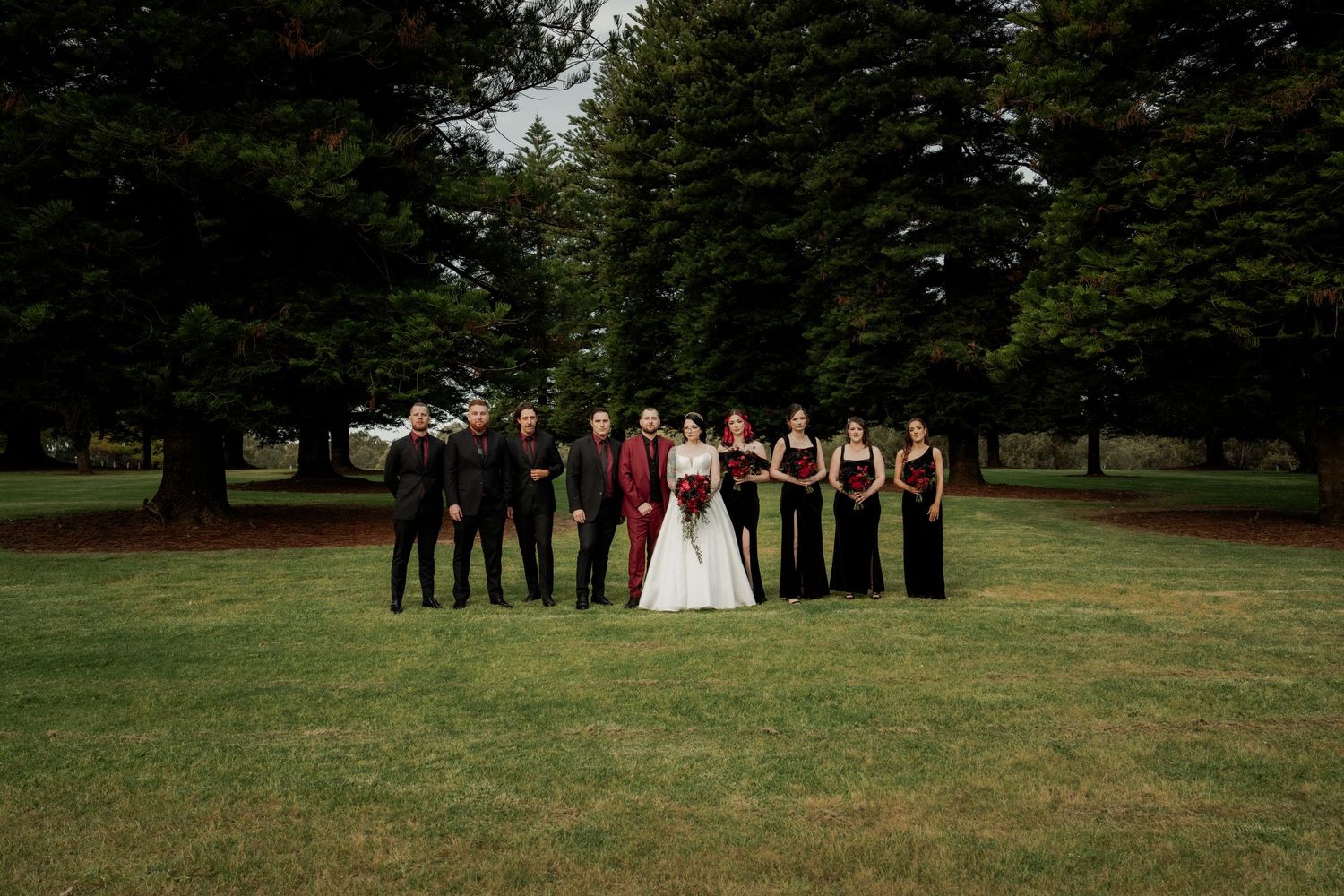 Wedding party in formal attire posing on green lawn surrounded by tall trees, bride in white gown with groomsmen and bridesmaids.