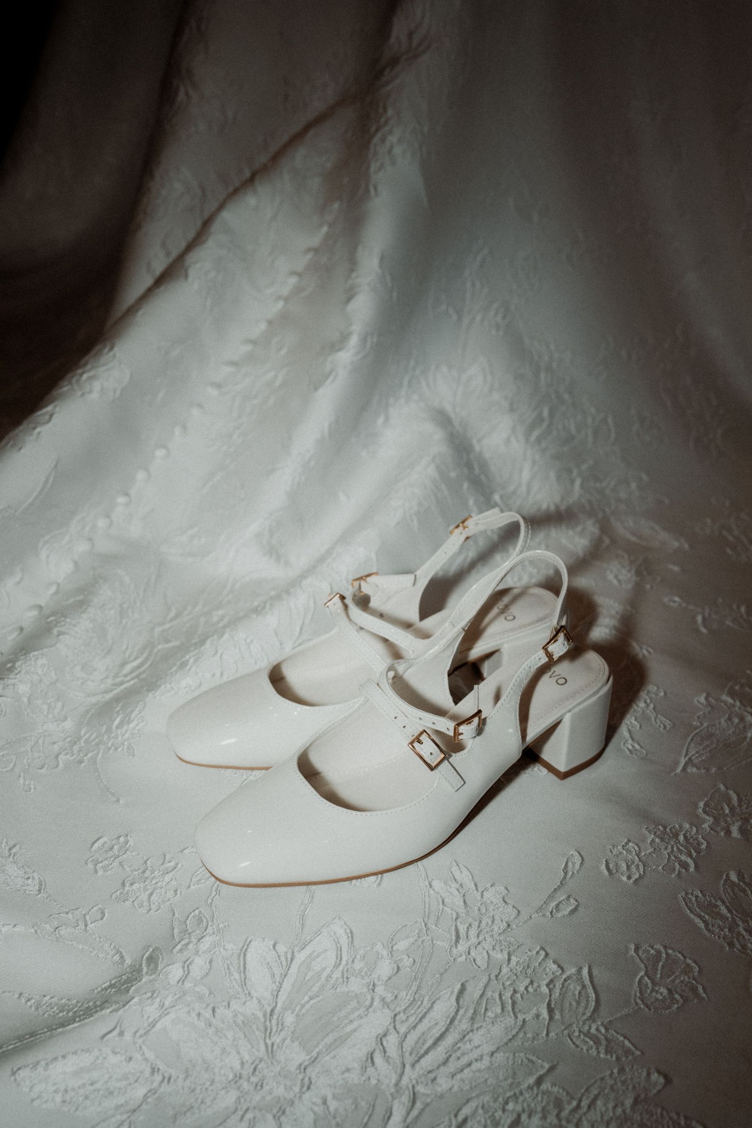 White block heel bridal shoes with double buckle straps displayed on an embossed white fabric background.