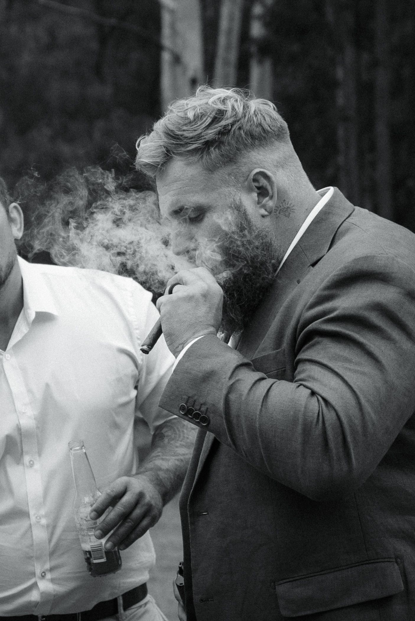 Black and white photo of a bearded man in a suit smoking a cigar outdoors at a formal event.