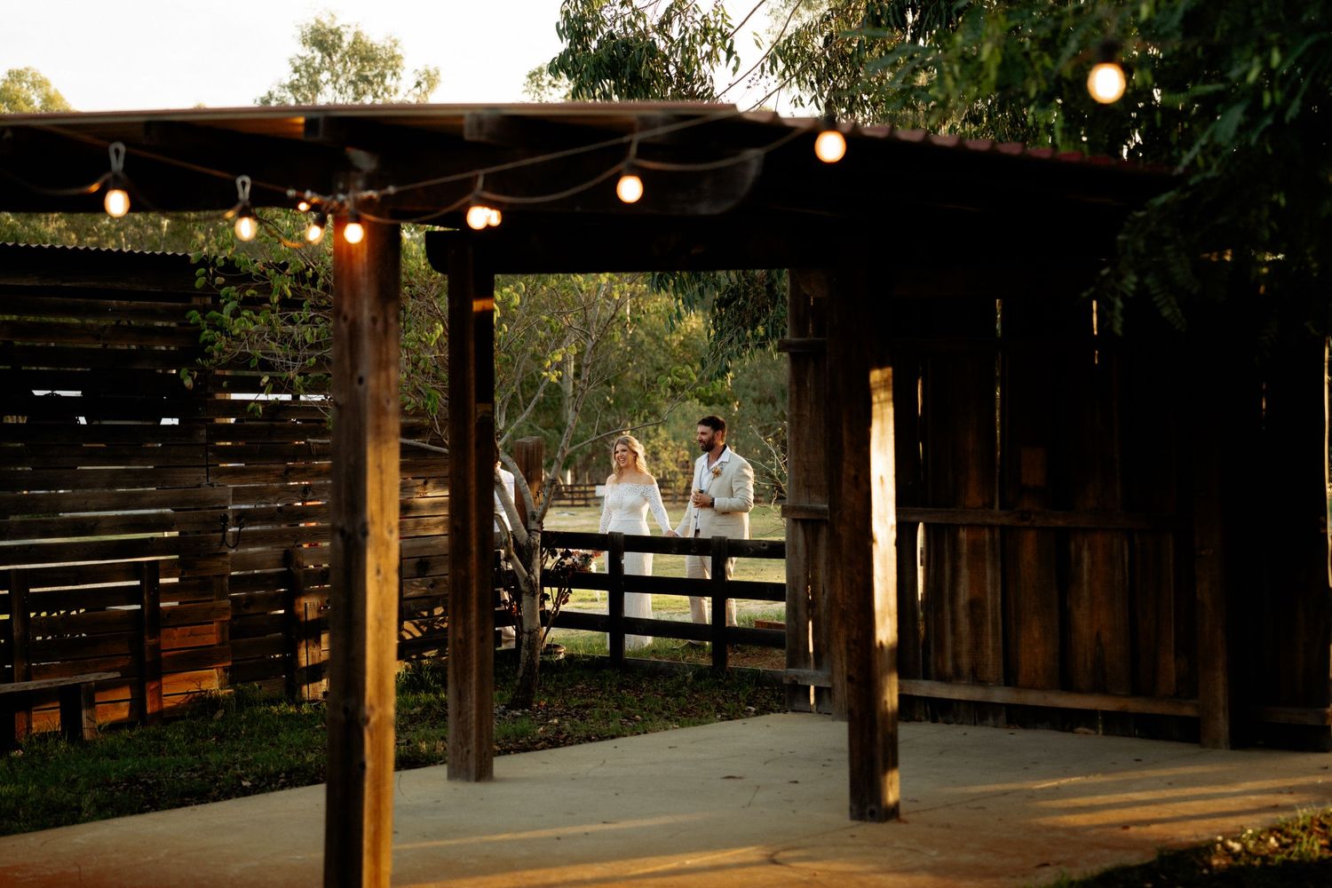 Bride and groom walking hand in hand under a rustic wooden pergola with string lights at dusk.