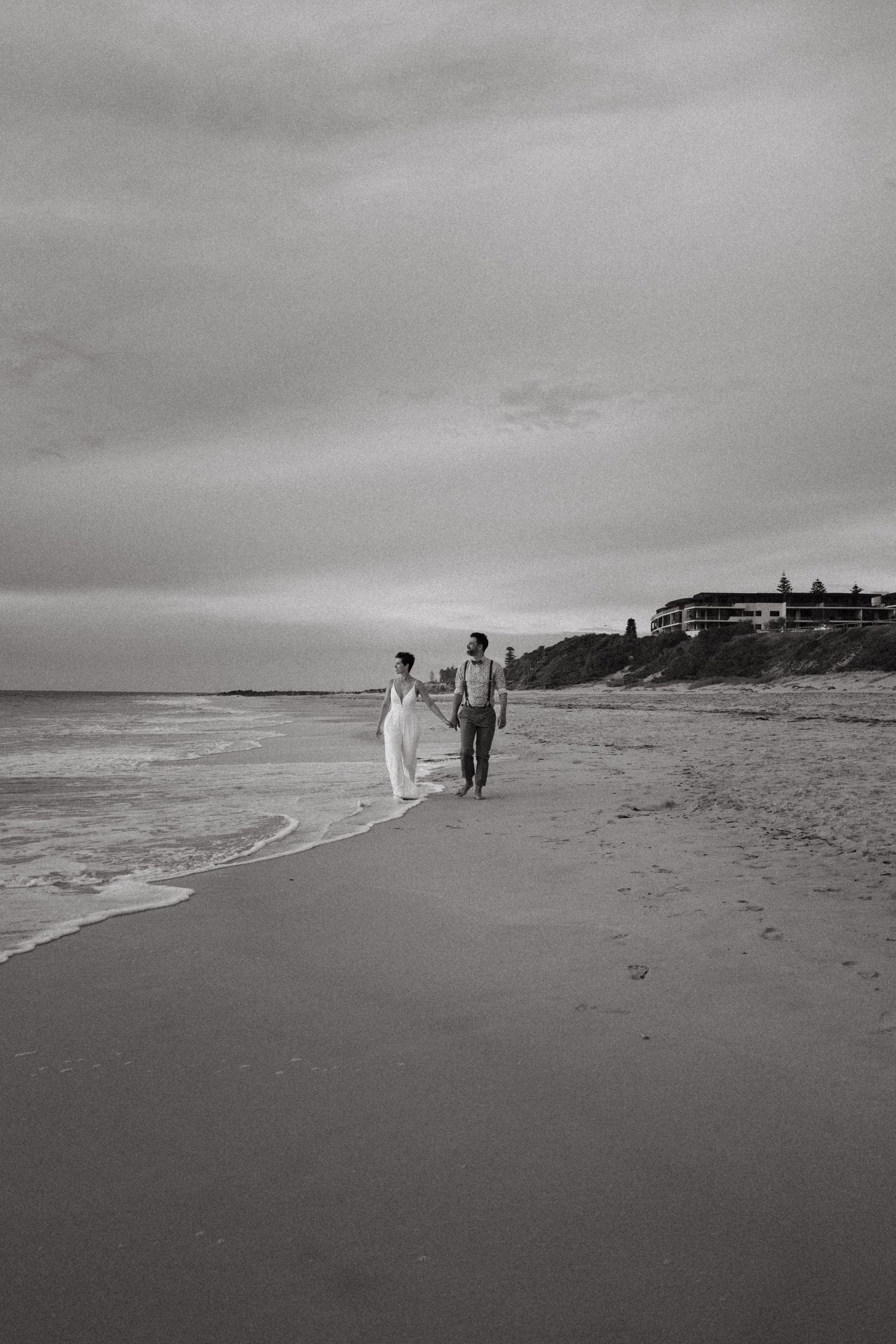 Black and white photo of a couple holding hands walking along a beach shoreline on a cloudy day.