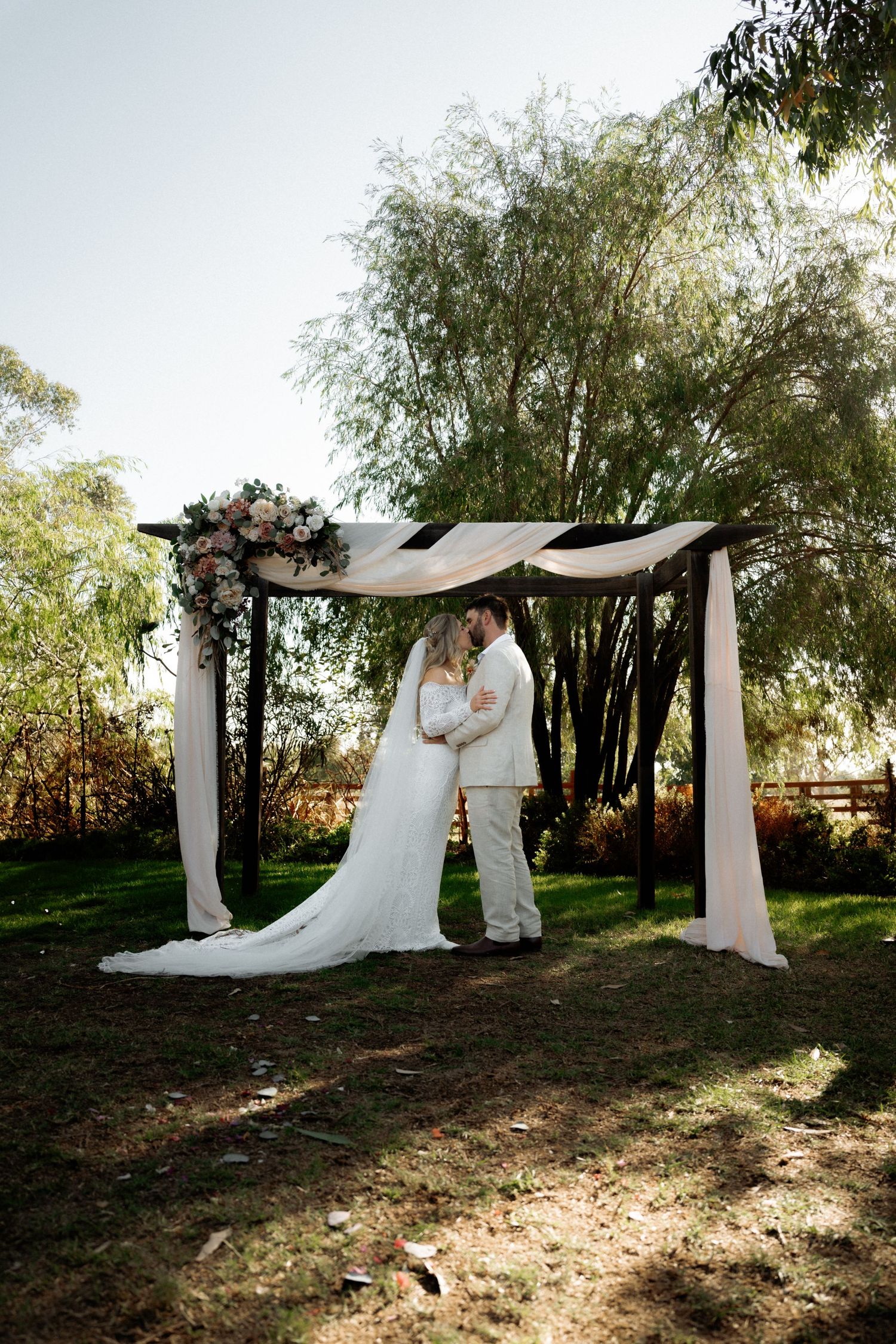 Bride and groom share a romantic kiss under a floral wedding arch draped with white fabric outdoors.